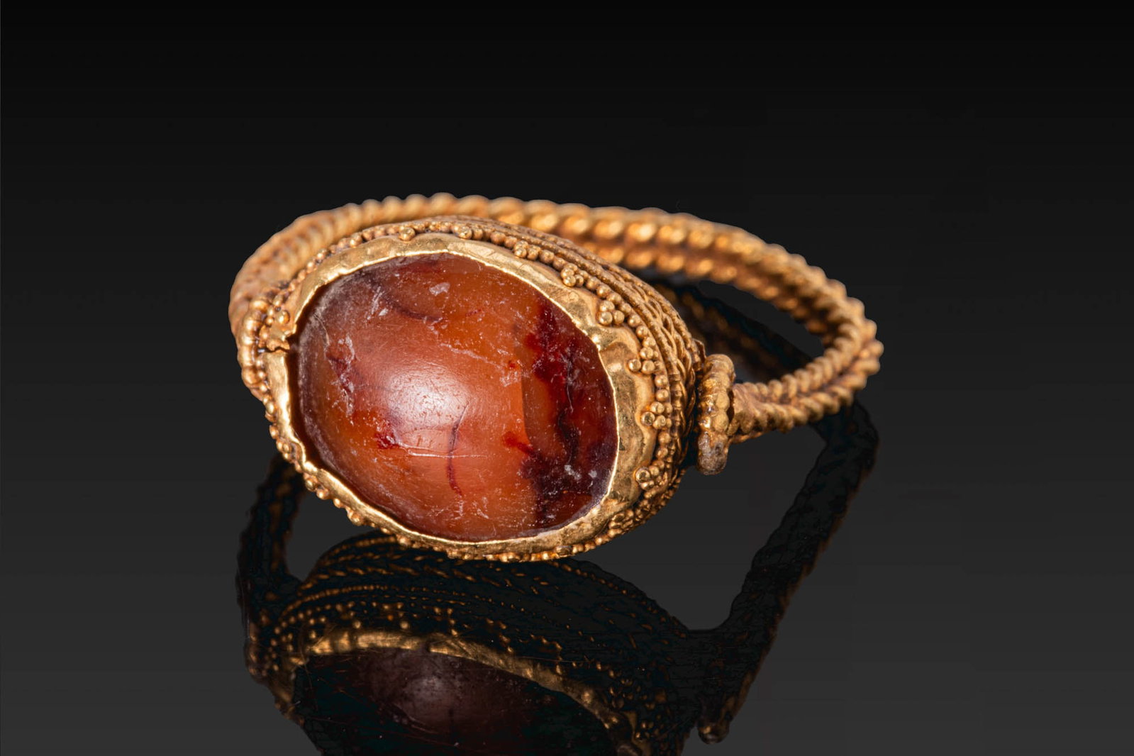 HELLENISTIC GOLD RING WITH CARNELIAN CABOCHON AND GRANULATION: Ca. 300 BC. A gold ring with a braided hoop and an oval bezel set with a carnelian cabochon. The setting is decorated with bands of twisted and beaded wire and clusters of applied granules. For simila