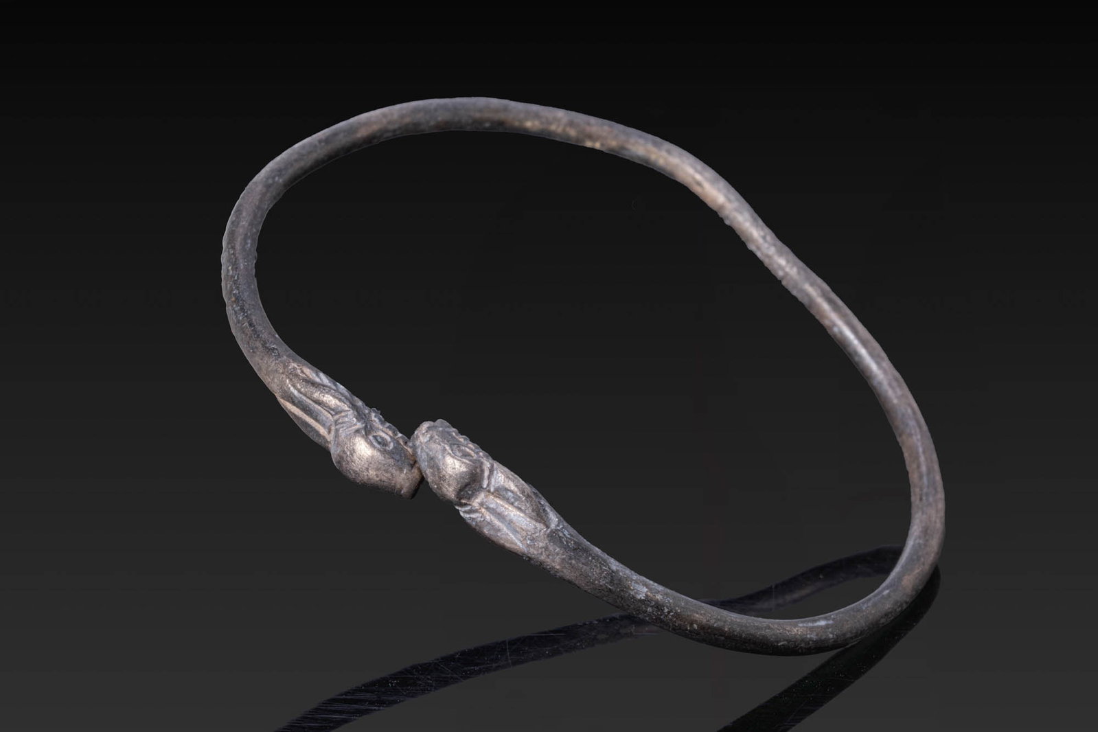 GREEK SILVER BRACELET WITH ZOOMORPHIC FINIALS: Ca. 500 BC. A silver bracelet formed from a slender round-section hoop with opposed zoomorphic terminals, each modelled as a stylised animal head with a narrow muzzle and incised eyes. For similar see