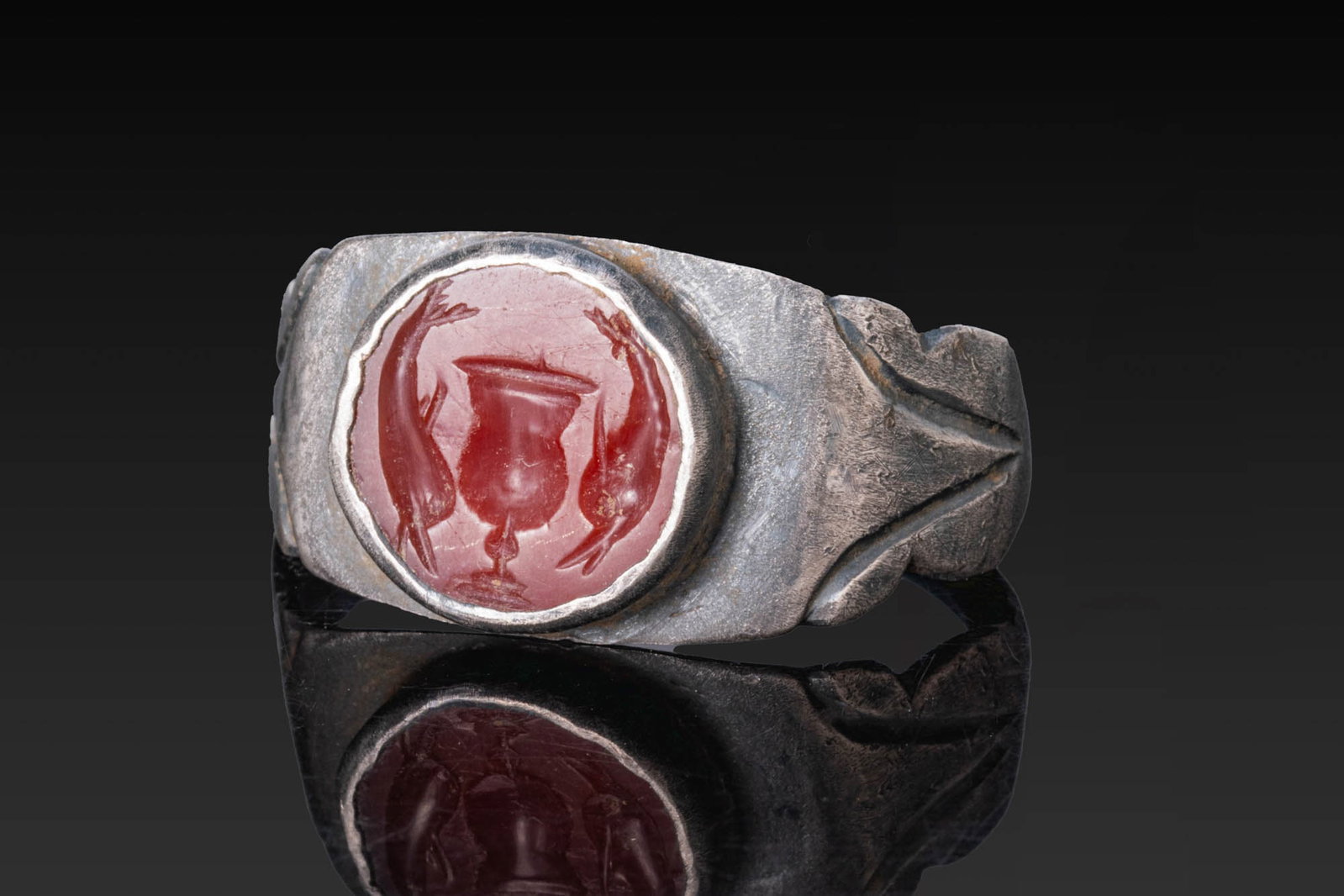 ROMAN CARNELIAN INTAGLIO OF A VASE FLANKED BY DOLPHINS IN SILVER RING: Ca. AD 1 - 200. A carnelian intaglio engraved with a tall vase flanked by two dolphins, set in a possibly later silver ring with a flat-section hoop widening to the shoulders cut with V-shaped motifs,