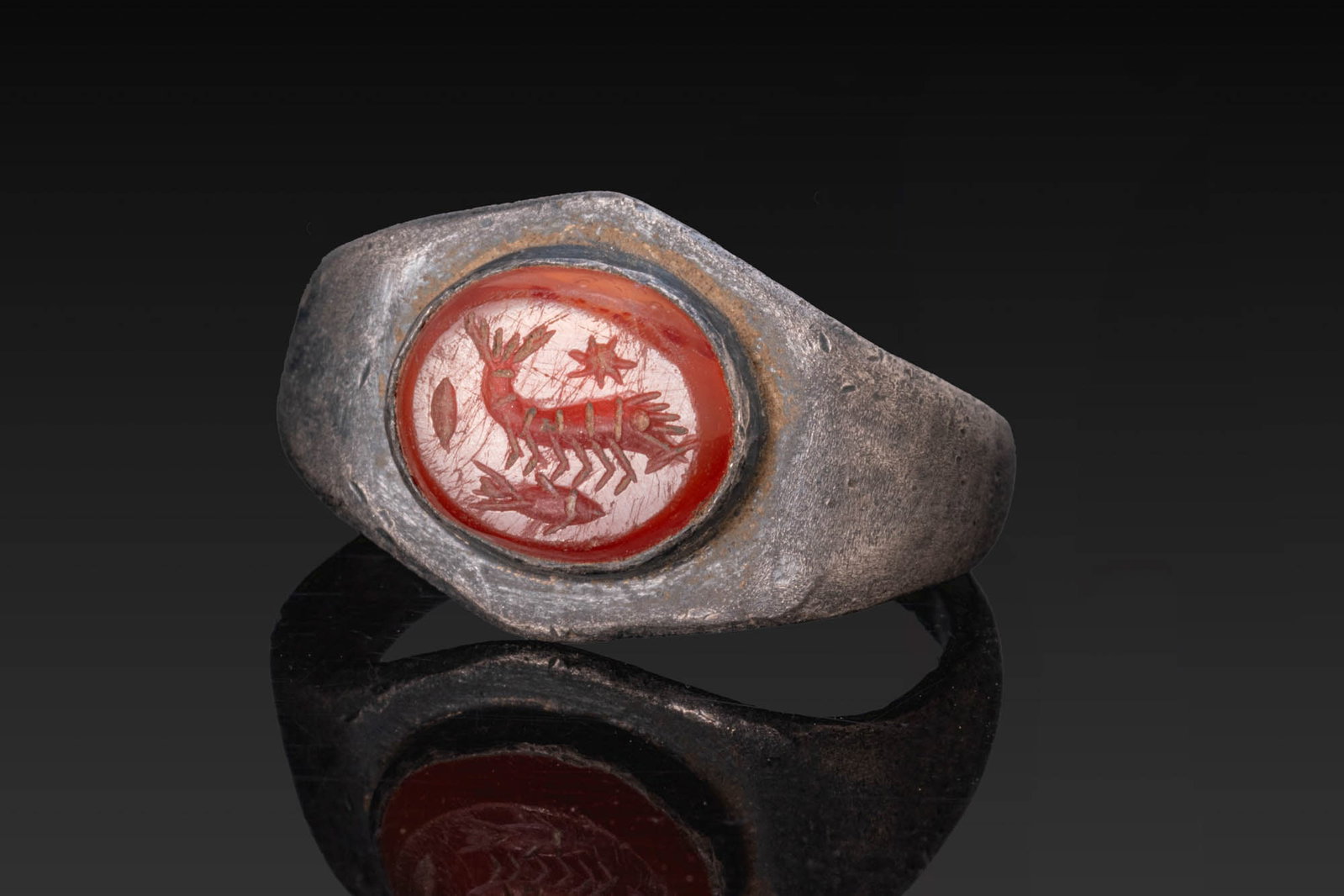 ROMAN CARNELIAN INTAGLIO OF SEA CREATURES IN SILVER RING: Ca. AD 1 - 300. A carnelian intaglio engraved with sea creatures, set in a possibly later silver ring with a flat-section hoop widening towards a hexagonal bezel plate with an oval cell. Size: D:18.34