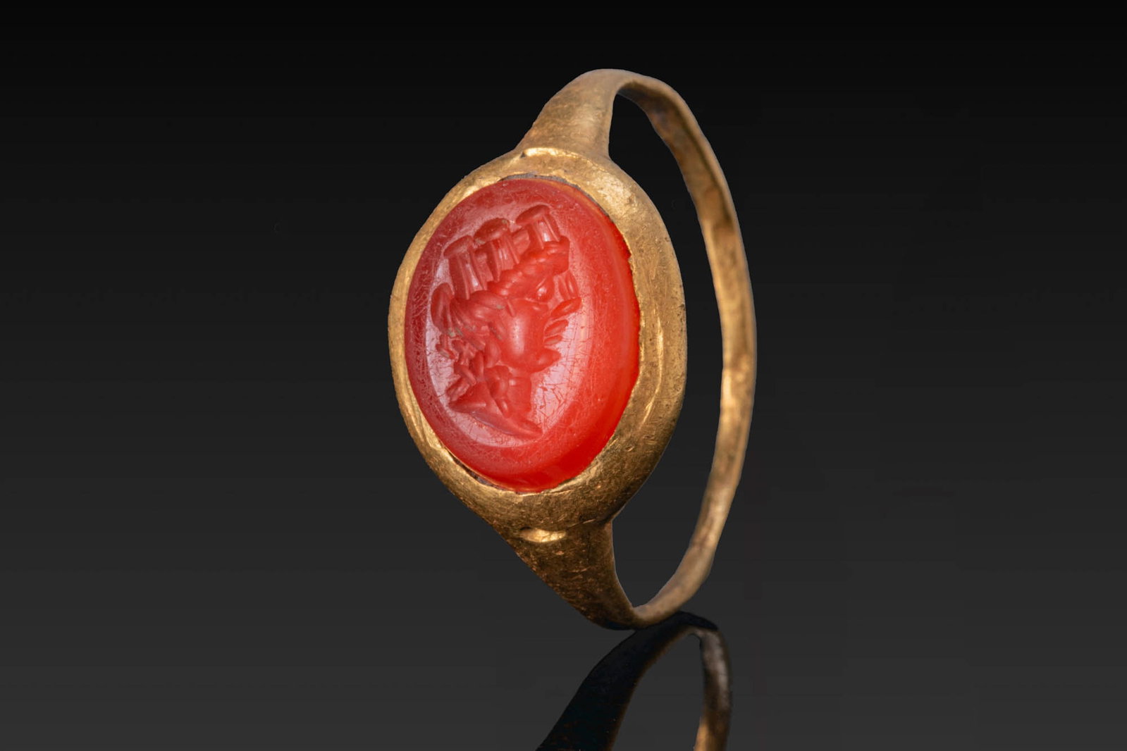 ROMAN GOLD RING WITH CARNELIAN INTAGLIO OF GODDESS FORTUNA: Ca. AD 300. A gold ring with a slender hoop broadening to an oval bezel set with a carnelian intaglio engraved with the profile portrait of Fortuna wearing a corona muralis. Size: D:15.9mm / US: 5 1/4