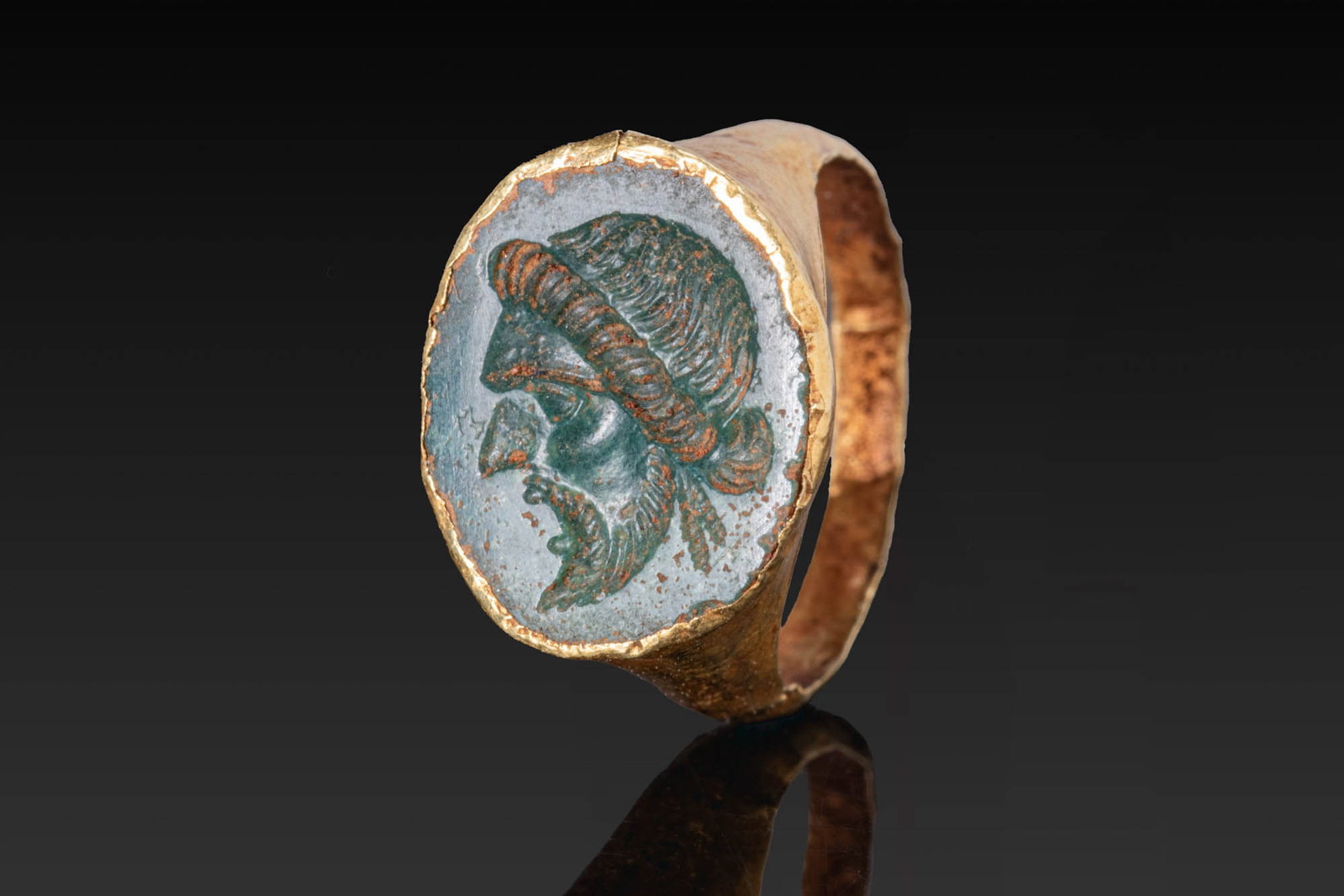 ROMAN GOLD RING WITH GLASS INTAGLIO OF JUPITER: Ca. AD 1 - 100. A gold ring with a rounded hoop and a tall oval bezel set with a green glass intaglio carved with the profile head of Jupiter, bearded and wearing a fillet. For similar see: The Classi
