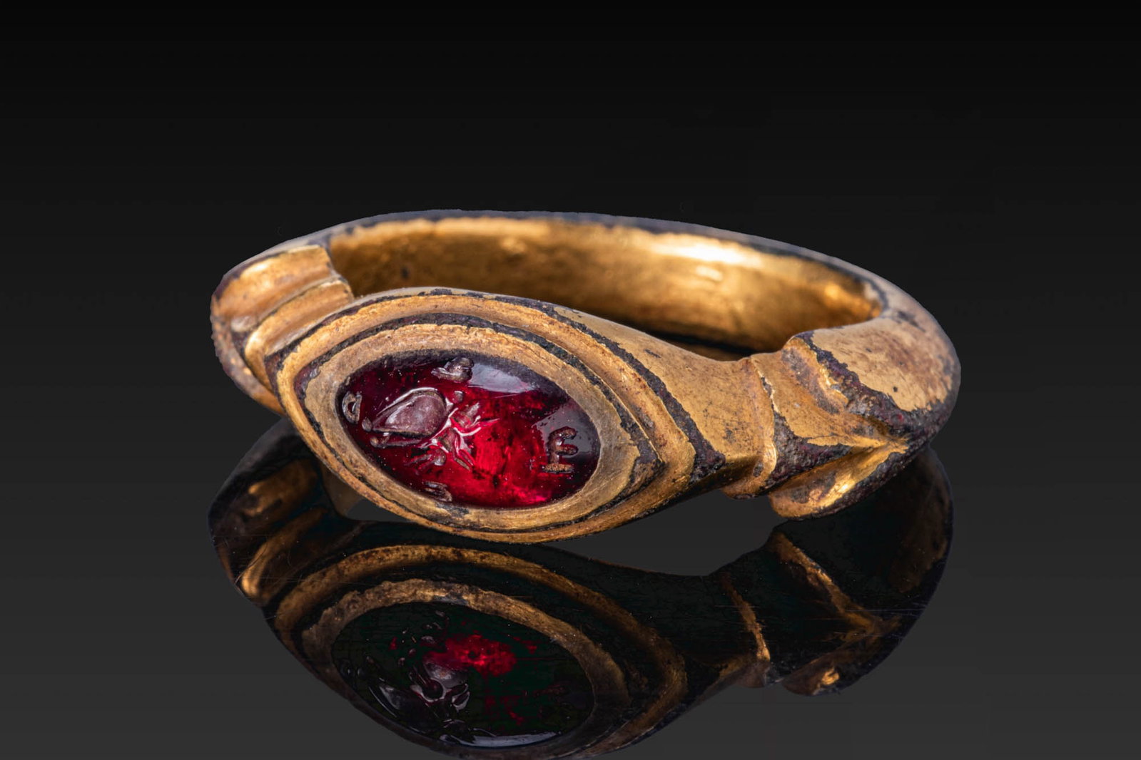 EASTERN ROMAN GILT-BRONZE RING WITH GARNET INTAGLIO OF A BUTTERFLY AND LETTERS: Ca. AD 300 - 500. A gilt-bronze ring with a carinated hoop, stepped shoulders and an elongated navette-shaped bezel with tiered borders, set with a garnet intaglio engraved with a butterfly framed by 