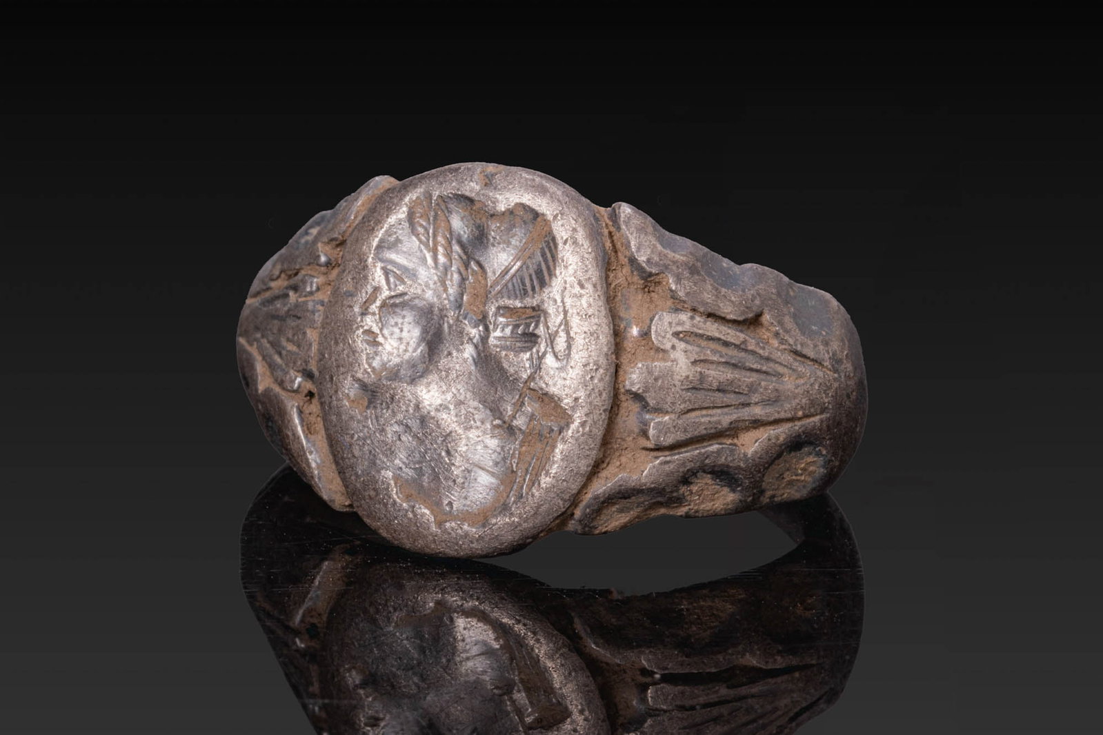 LATE ROMAN SILVER RING WITH NOBLEWOMAN: Ca. AD 200 - 400. A silver ring with a broad hoop, expanding to slightly angular shoulders decorated with vegetal motifs, and an oval bezel engraved with the profile bust of a noblewoman. Size: D:19.1