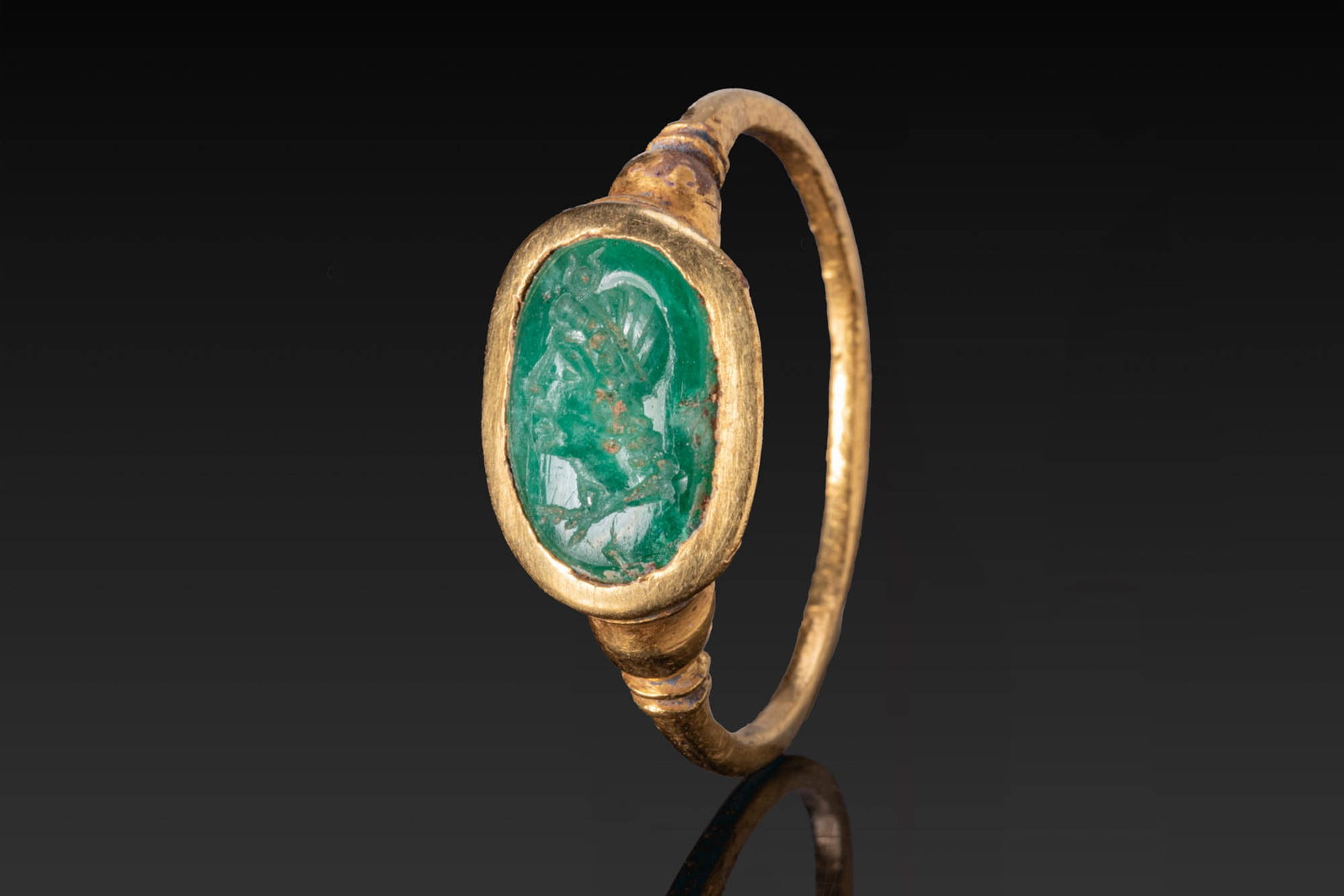 ROMAN GOLD RING WITH EMERALD INTAGLIO OF A PROFILE PORTRAIT OF ISIS: Ca. AD 1 - 200. A gold ring with a slender hoop, stepped shoulders and an oval bezel set with an emerald intaglio engraved with the profile portrait of Isis wearing a diadem and horned crown. For simi