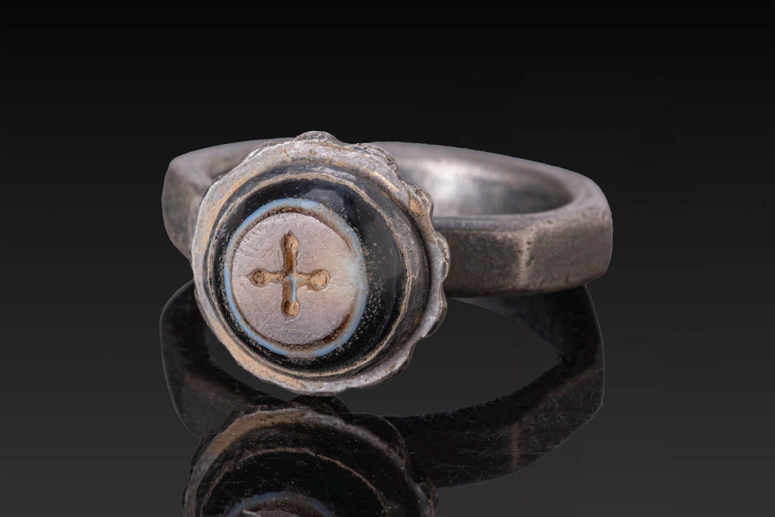 BYZANTINE AGATE INTAGLIO OF A CROSS IN SILVER RING: Ca. AD 600. A convex agate intaglio engraved with a cross with rounded finials, set in a silver ring with a polygonal flat-section hoop and a cup-shaped bezel with a fluted underside and a scalloped o