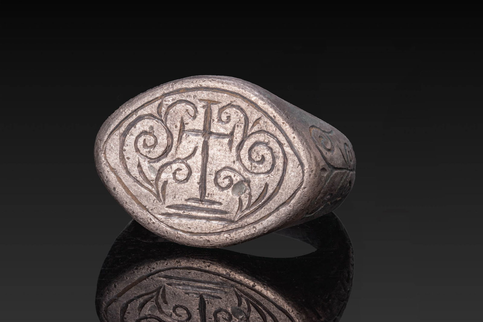 BYZANTINE SILVER RING WITH CROSS FLANKED BY VEGETAL TENDRILS: Ca. AD 1200 - 1400. A silver ring with a broad D-shaped hoop widening towards a large slightly pointed oval bezel engraved with a cross on a stepped base, flanked by spiral motifs. The shoulders are e