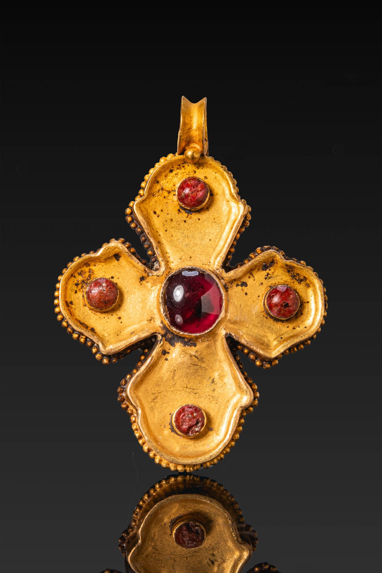 BYZANTINE GOLD CROSS PENDANT WITH GARNET AND GLASS CABOCHONS: Ca. AD 600 - 800 (or later). A gold cross pendant with broad lobed arms, each set with a red glass cabochon, centred by a garnet, with a plain suspension loop above. The outer edge is bordered with gr