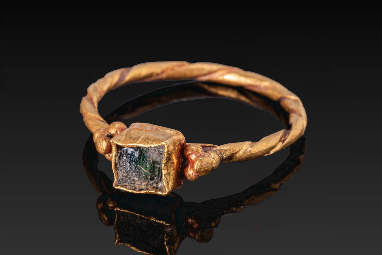 EARLY BYZANTINE GOLD TWISTED RING WITH GLASS CABOCHON: Ca. AD 300 - 500. A gold ring with a twisted hoop and a square bezel setting with granulated shoulders and a green glass cabochon. Size: D:17.12mm / US: 6 3/4 / UK: N; Weight: 2.7g Provenance: Private