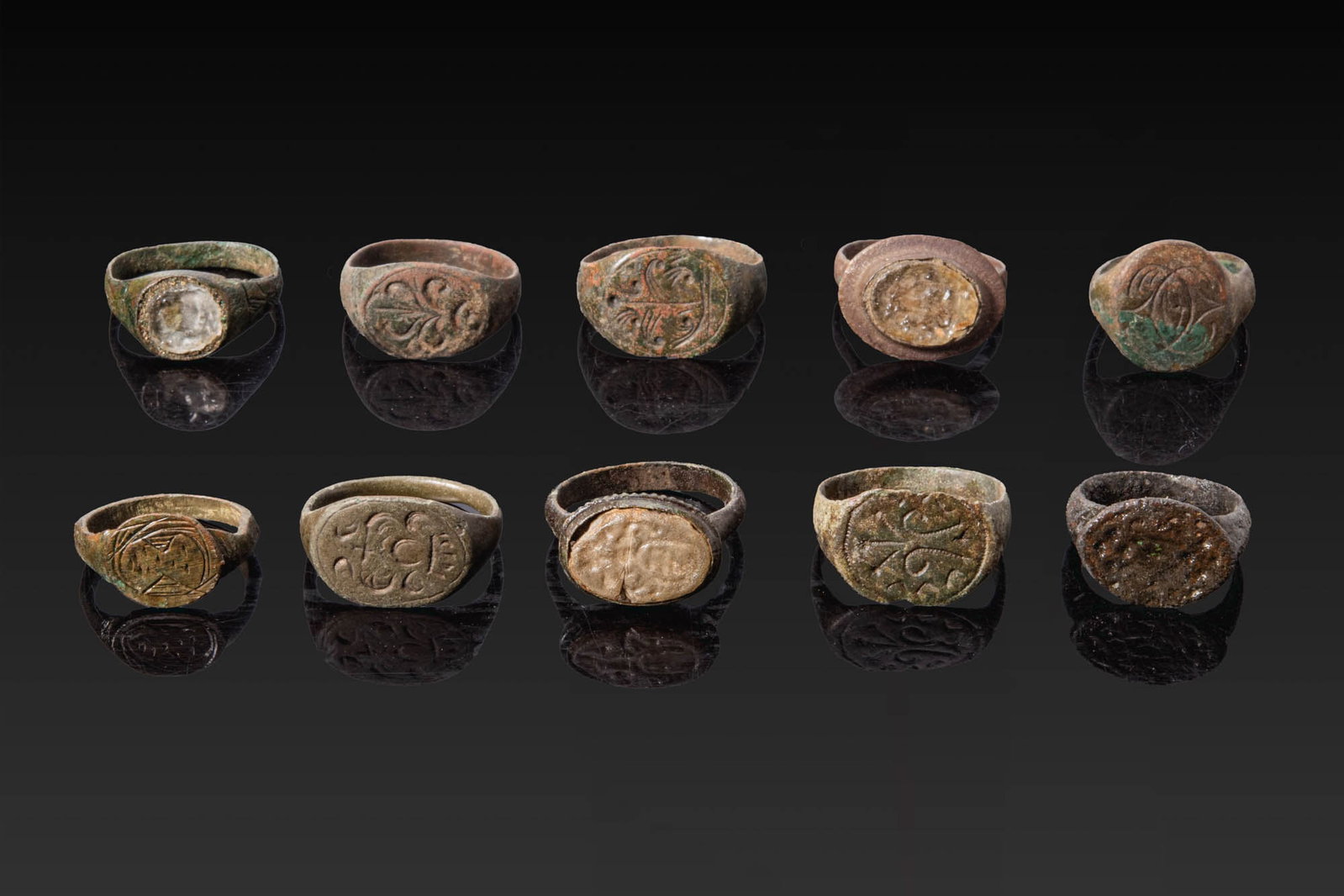 COLLECTION OF MEDIEVAL BRONZE RINGS WITH HERALDIC SYMBOLS, GEOMETRIC MOTIFS AND GLASS GEMS: Ca. AD 1000 - 1300. A collection of ten bronze rings, including, from top left: a ring with a round bezel set with a pale glass inset; a ring with an oval bezel engraved with a quatrefoil device; a ri