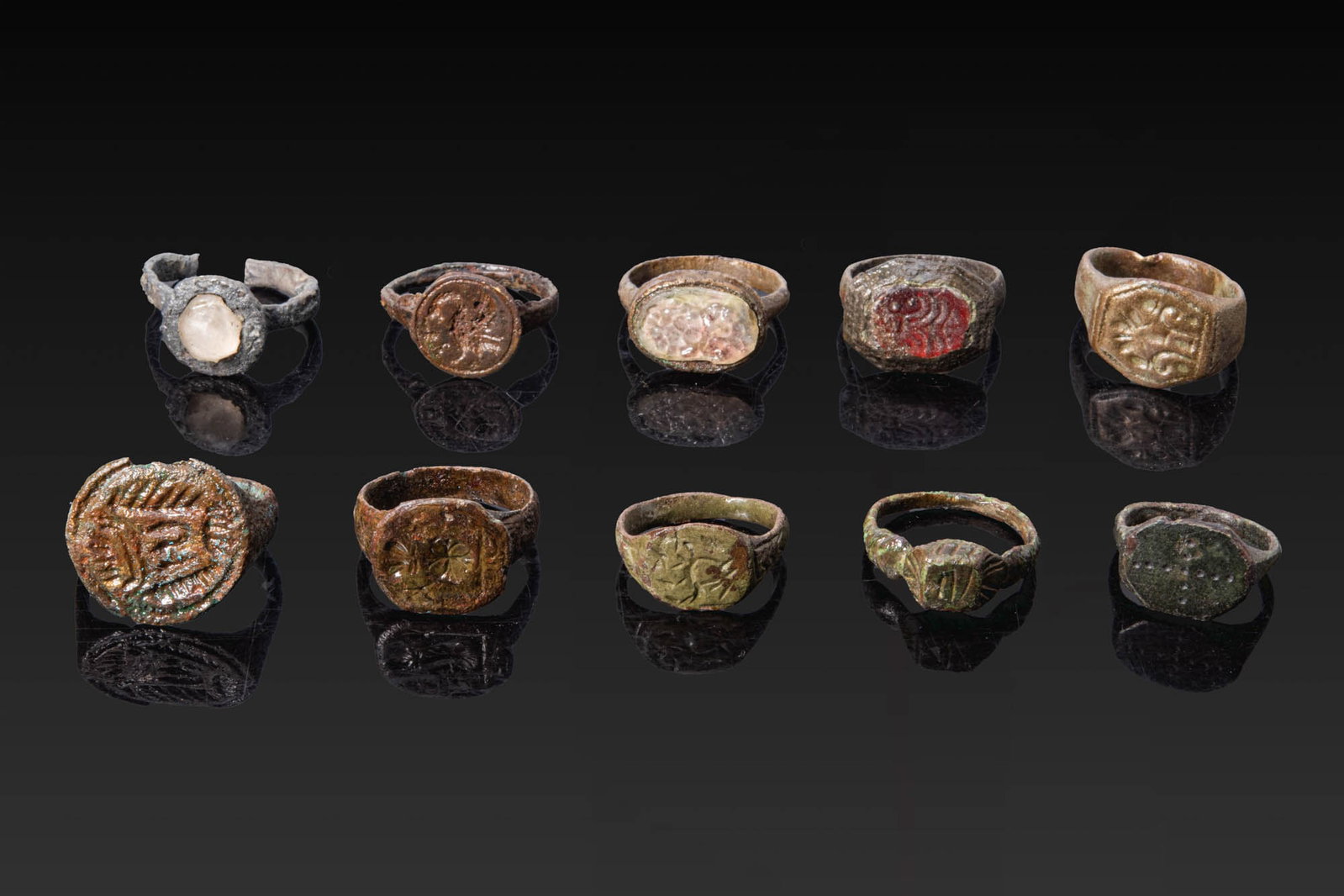 COLLECTION OF MEDIEVAL BRONZE RINGS WITH HERALDIC SYMBOLS, GEOMETRIC MOTIFS AND GLASS GEMS: Ca. AD 1000 - 1300. A collection of ten bronze rings, including, from top left: a ring with a round bezel set with a pale glass inset; a ring with a round bezel engraved with a bird in profile; a ring