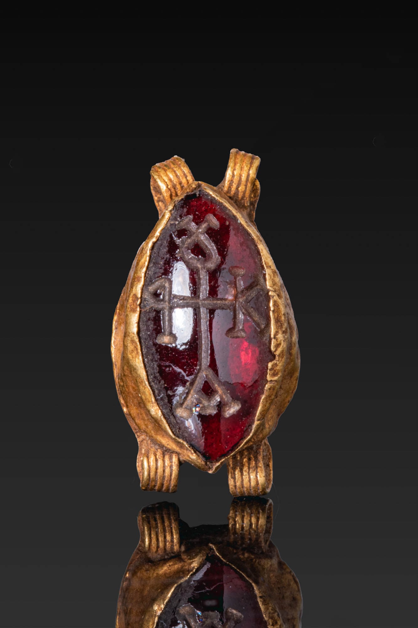 BYZANTINE OR MEROVINGIAN GOLD AND GARNET PENDANT WITH MONOGRAM: Ca. AD 500 - 700. A gold pendant with an elongated garnet cut en cabochon, engraved with a cruciform monogram with Greek letters at the ends of the arms. The stone is set in a plain high cell, with pa
