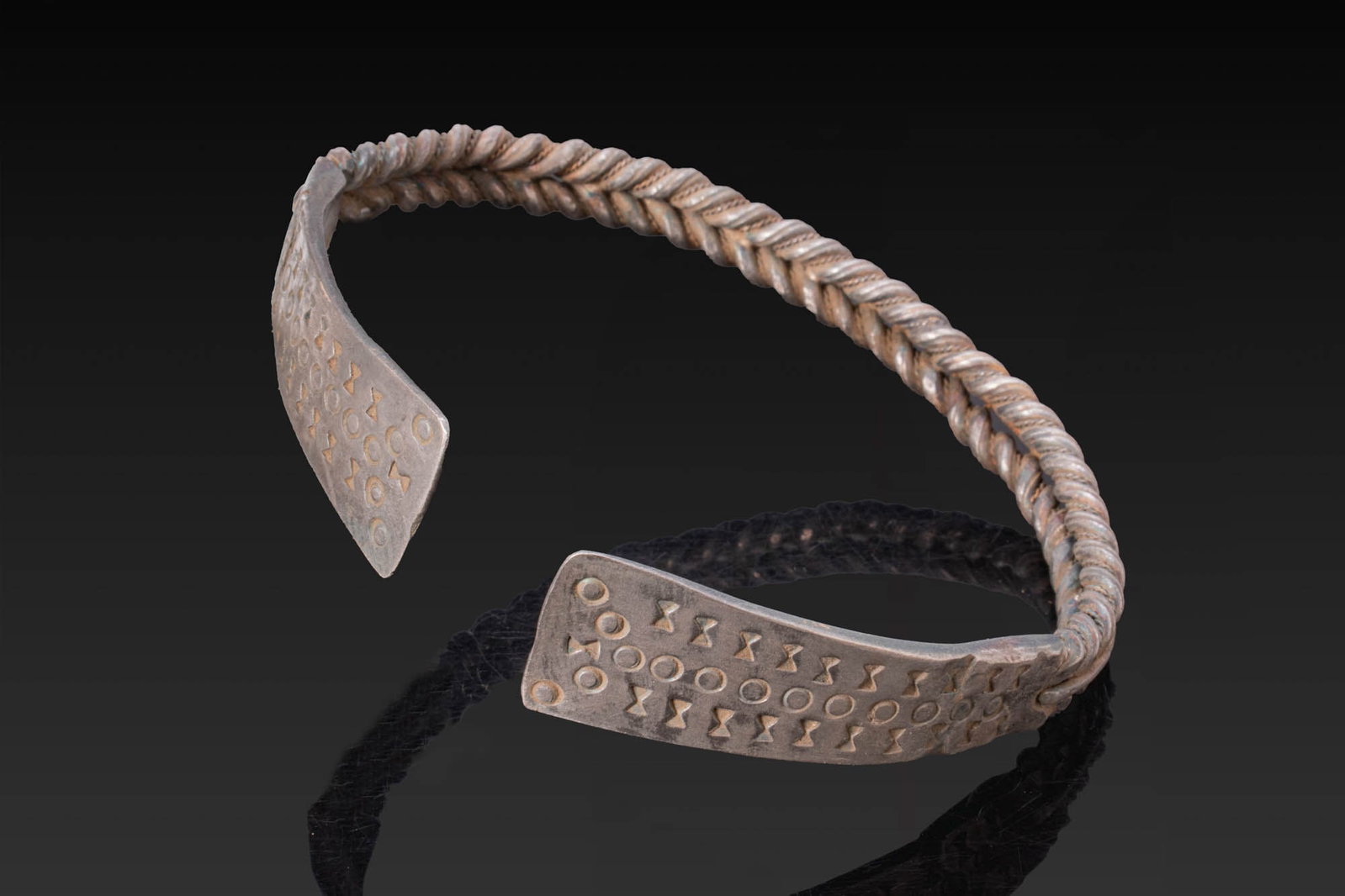 VIKING SILVER BRAIDED BRACELET WITH STAMPED FINIALS: Ca. AD 800 - 1000. A silver penannular bracelet with a braided shank widening at each end into flat flaring finials decorated with stamped rings and hourglass-shaped impressions arranged in horizontal
