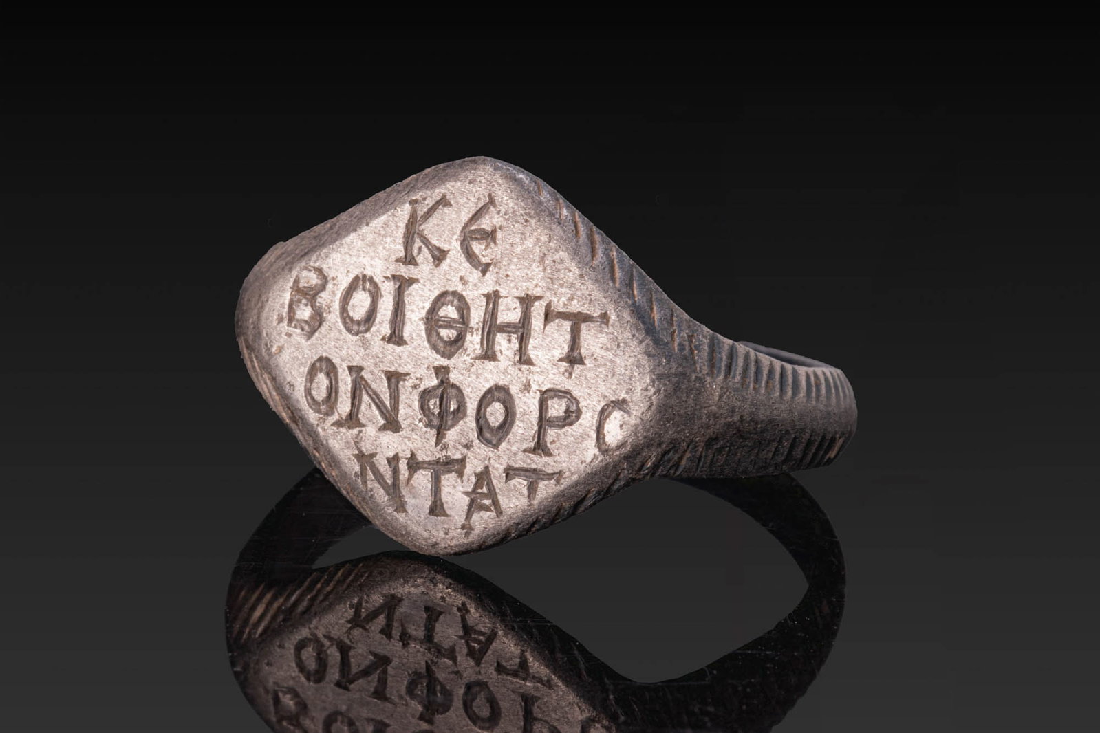 BYZANTINE SILVER RING WITH GREEK INSCRIPTION: Ca. AD 900 - 1100. A silver ring with a lozenge-shaped bezel engraved with a four-line Greek inscription reading "?? / ?????? / ?????C / ????". The hoop tapers from the bezel and is decorated on the e