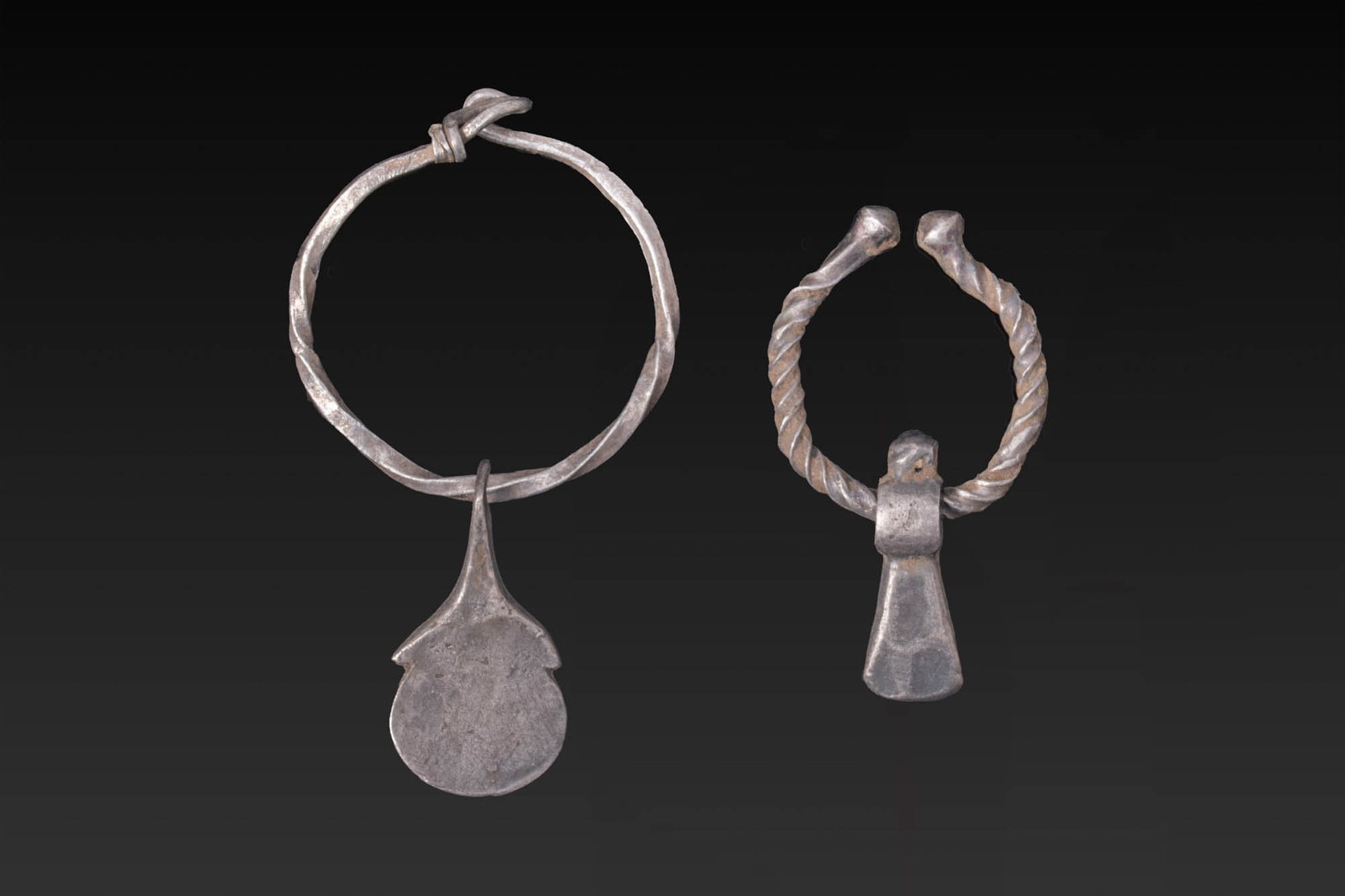 PAIR OF VIKING SILVER AXE-SHAPED PENDANTS: Ca. AD 700 - 1100. A pair of silver axe-shaped pendants, from left to right: a pendant with a flat circular blade and a tapering neck with pointed shoulders, suspended from a plain round-section ring 