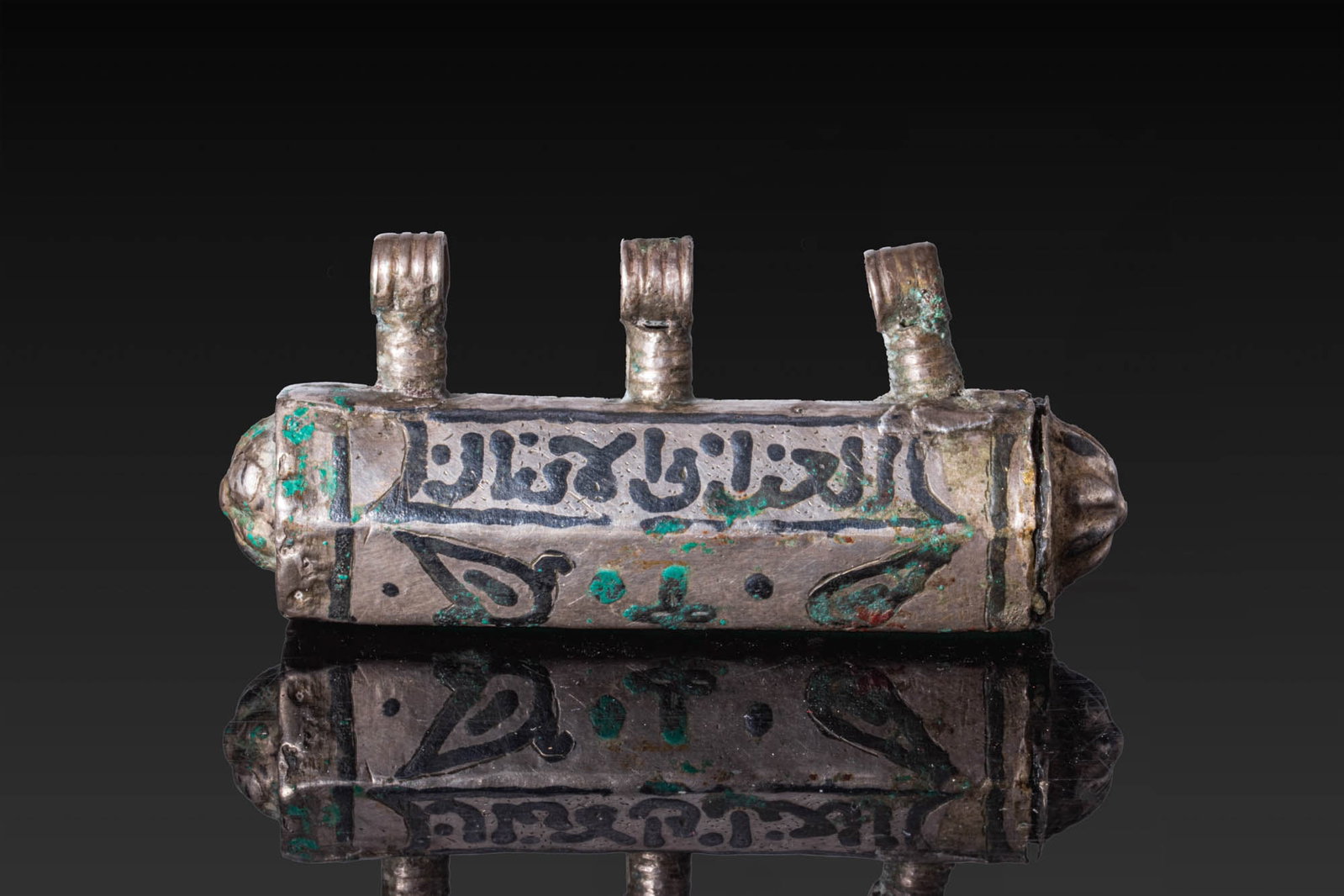 SELJUK SILVER AND NIELLO SCROLL PENDANT: Ca. AD 1000 - 1200. A silver scroll pendant formed as a tube of hexagonal section, with repousse rosettes on the domed terminals and three ribbed suspension loops above. Both sides are decorated with 