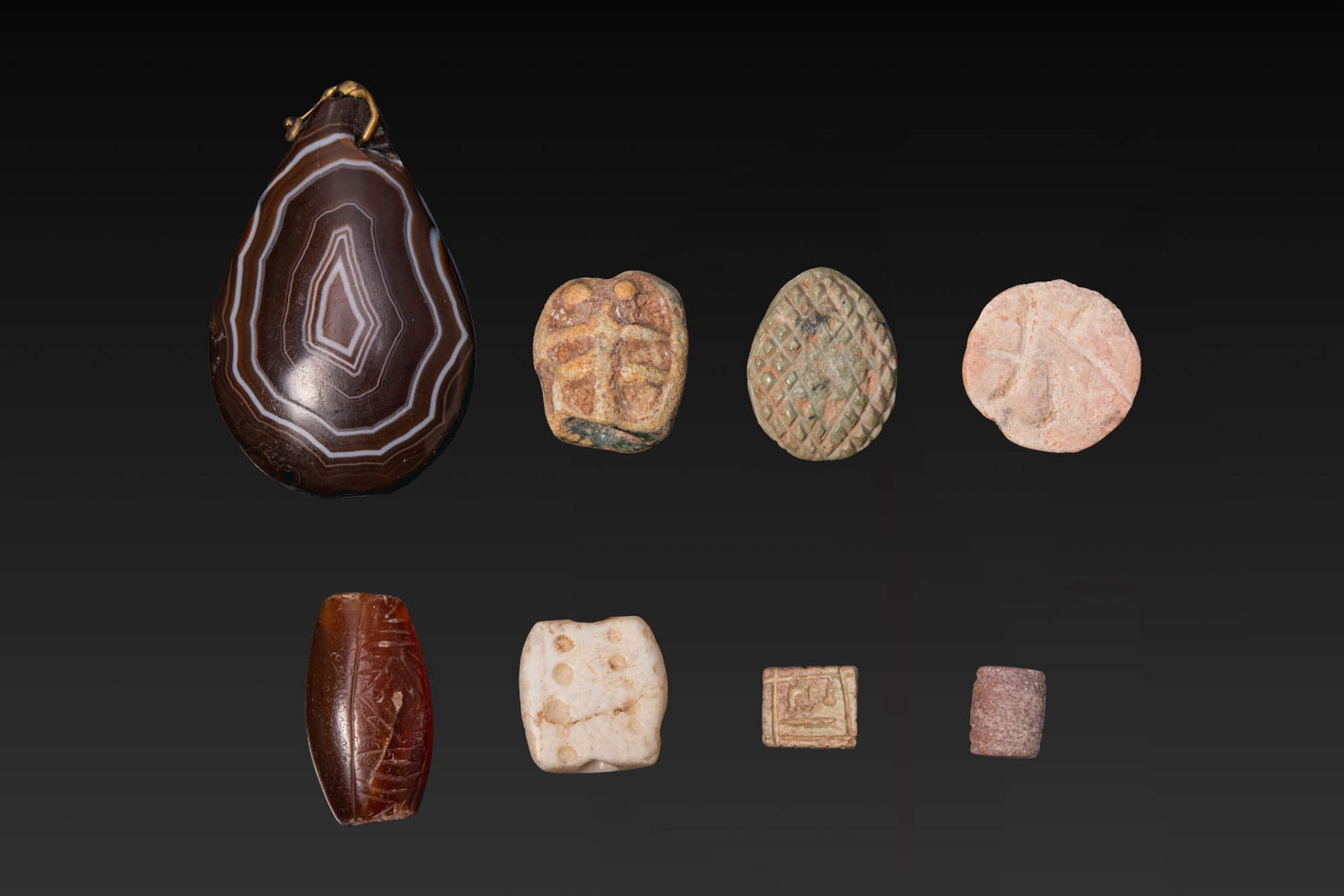 COLLECTION OF BACTRIAN STONE AMULETS, STAMP SEALS, BEADS AND PENDANTS: Ca. 2nd - 1st millennium BC. A collection of eight stone amulets, beads, stamp seals and pendants, including, from left to right: a large teardrop-shaped agate pendant with a gold suspension loop; two