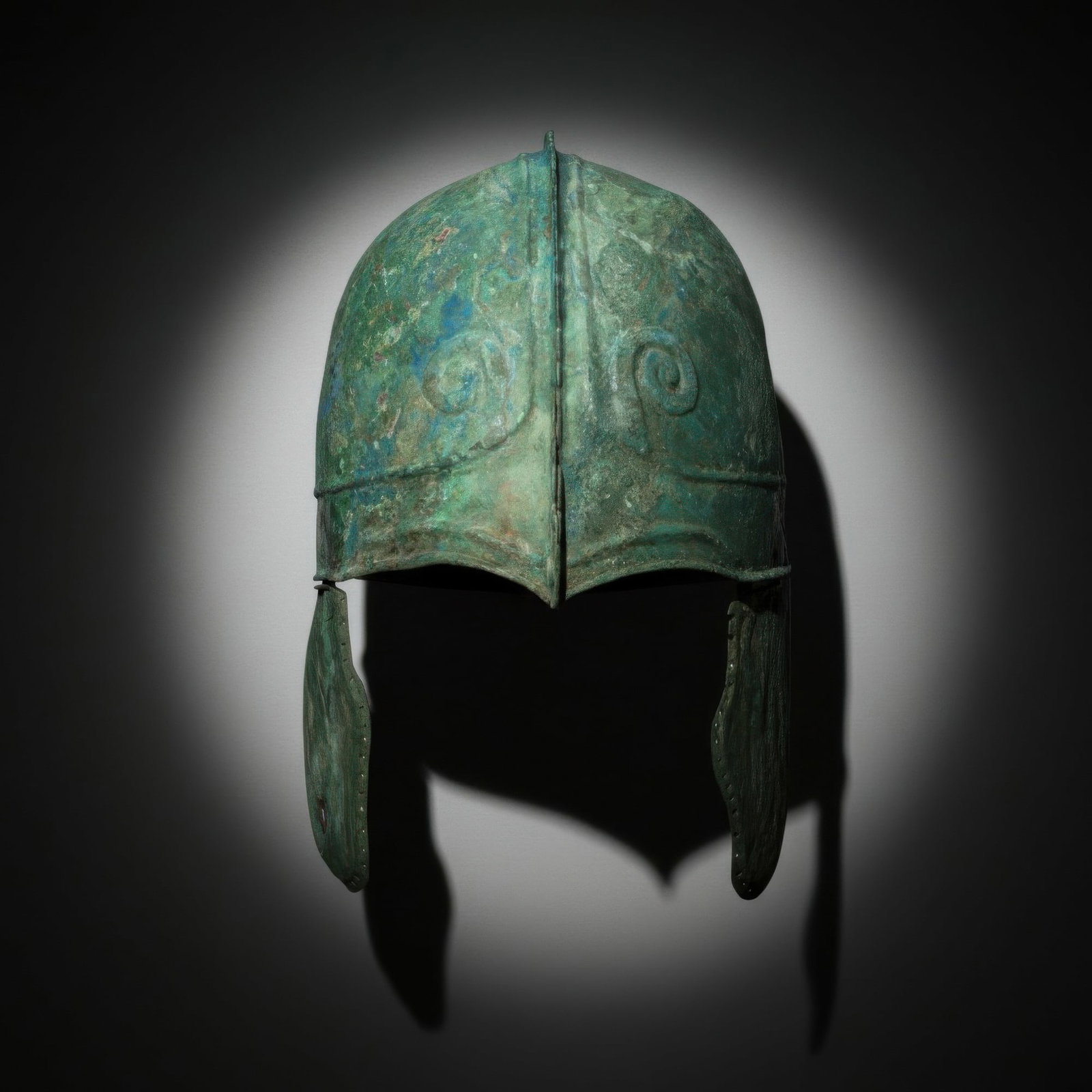 ANCIENT GREEK BRONZE HELMET WITH HORNS OF ZEUS AMUN AND CHEEK PIECES: Ca. 500 - 300 BC. A bronze helmet with a high rounded bowl, made of two halves riveted together at the centre and forming a short crest. A raised band runs around the lower edge, and the front is embo