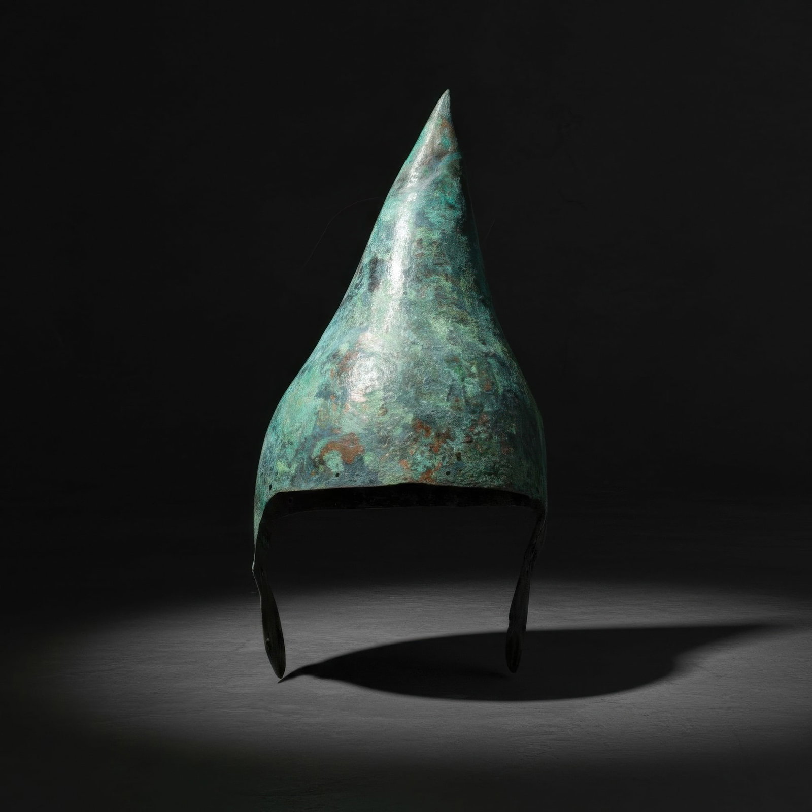 RARE ASSYRIAN BRONZE CONICAL HELMET WITH CHEEK PIECES: Ca. 800 - 600 BC. A bronze helmet with a tall conical crown rising to a pointed apex and two tongue-shaped cheekpieces pierced with attachment holes. For similar see: S. Borin, R. Sennewald, The Art o