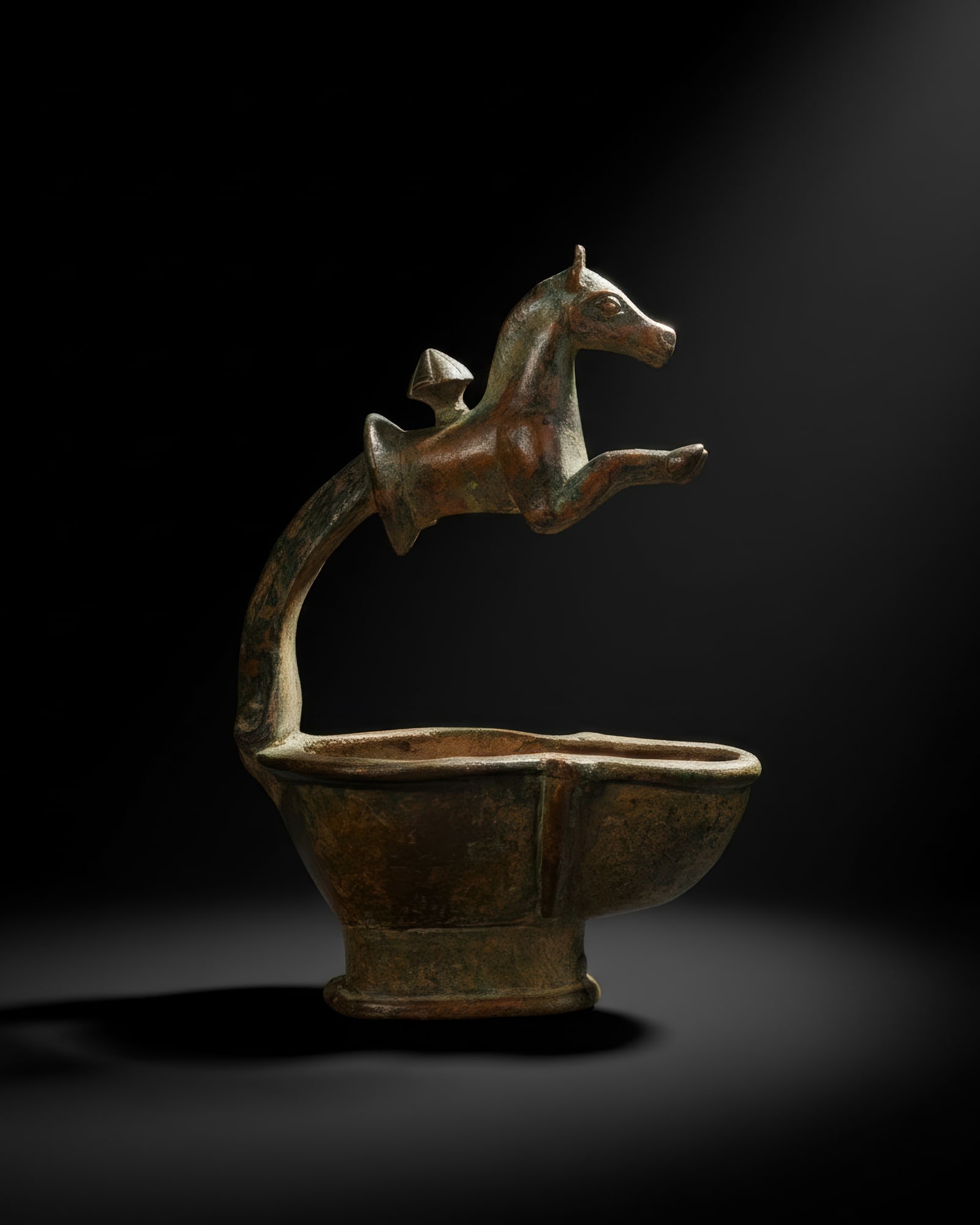 ROMANO-SABEAN BRONZE OIL LAMP WITH HORSE PROTOME HANDLE: Ca. AD 300 - 500. A bronze oil lamp with a deep body, a narrow raised rim, and a projecting nozzle. The arched handle is surmounted by the forepart of a horse, cast with extended forelegs, a long neck
