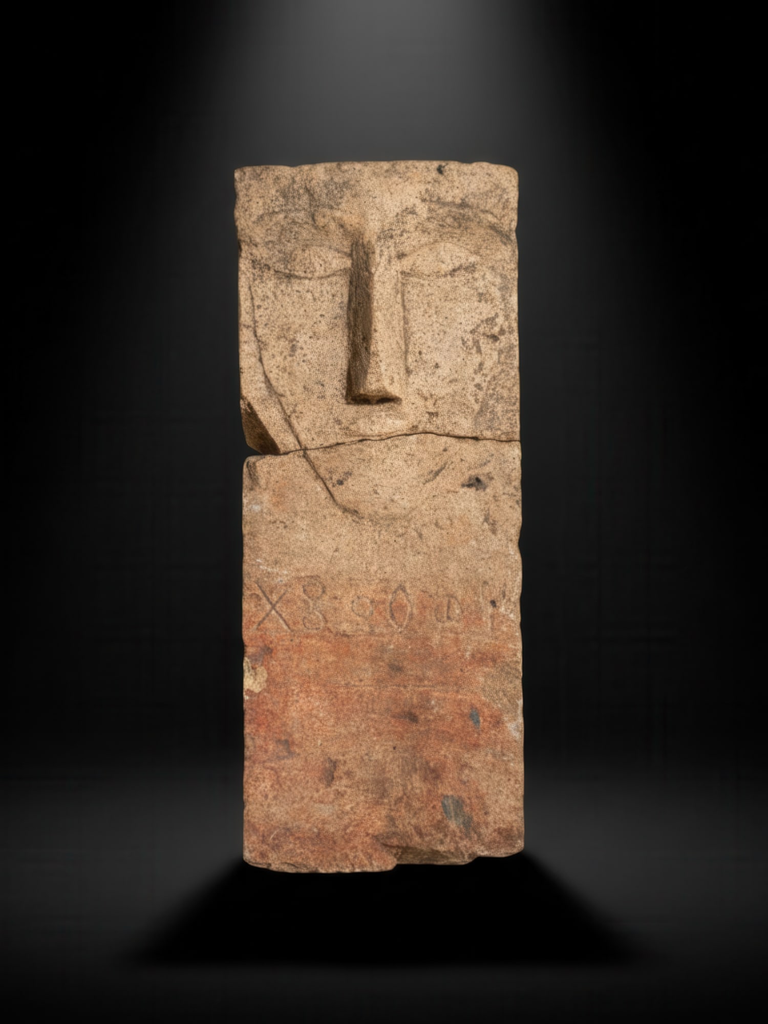 SOUTH ARABIAN INSCRIBED STONE FUNERARY STELE: Ca. 300 - 100 BC. A stone funerary stele with an abstract human face carved in shallow relief at the upper section. The face has arched brows, almond-shaped eyes, a long, straight nose, and a short, i