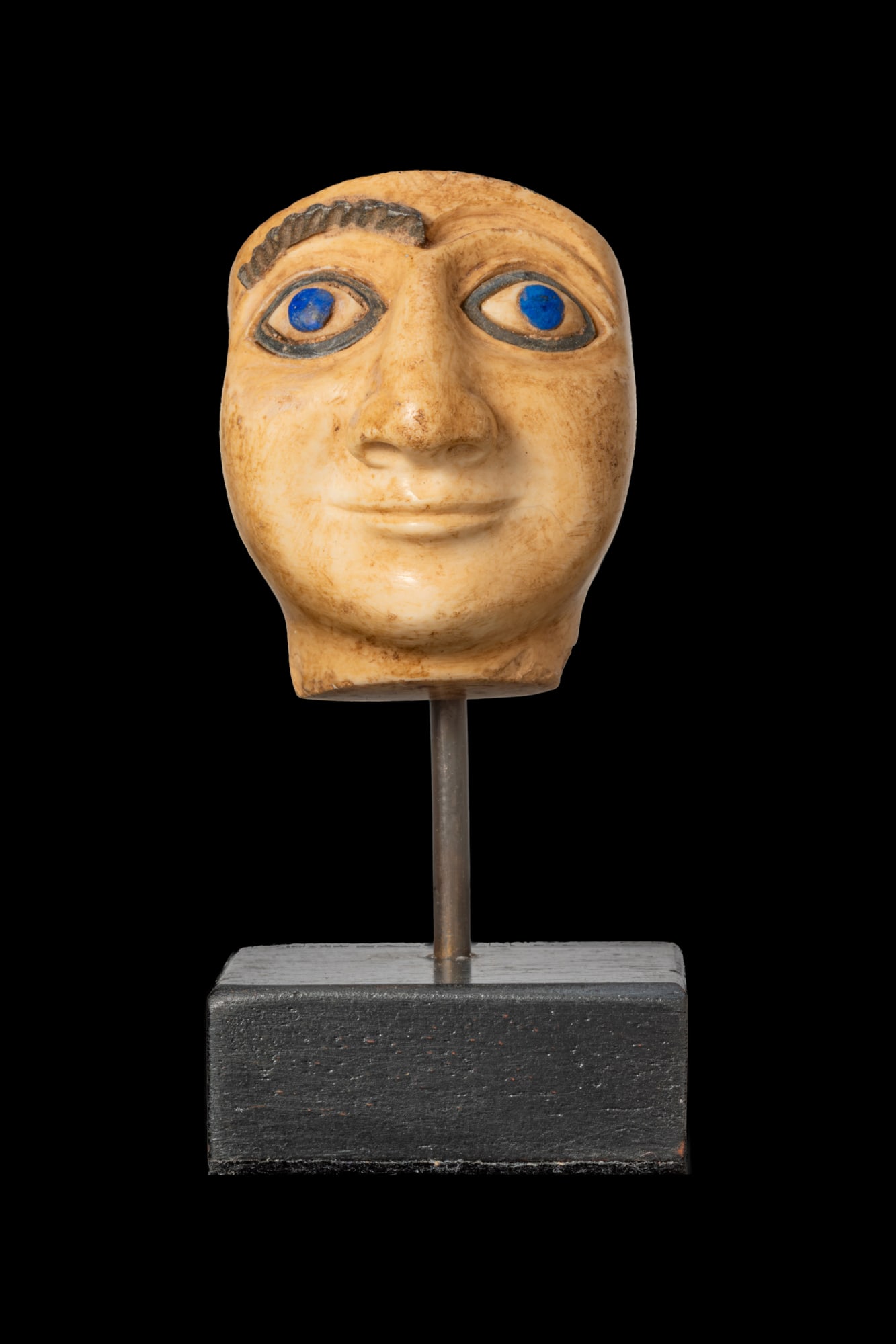 MESOPOTAMIAN STONE HEAD OF A WORSHIPPER WITH INLAID EYES AND BROW: Ca. 2600 - 2200 BC. A stone head of a worshipper with a broad face carved in the round. The eyes are inlaid with blue lapis lazuli pupils and white sclerae, set within dark stone rims. A ribbed brow i