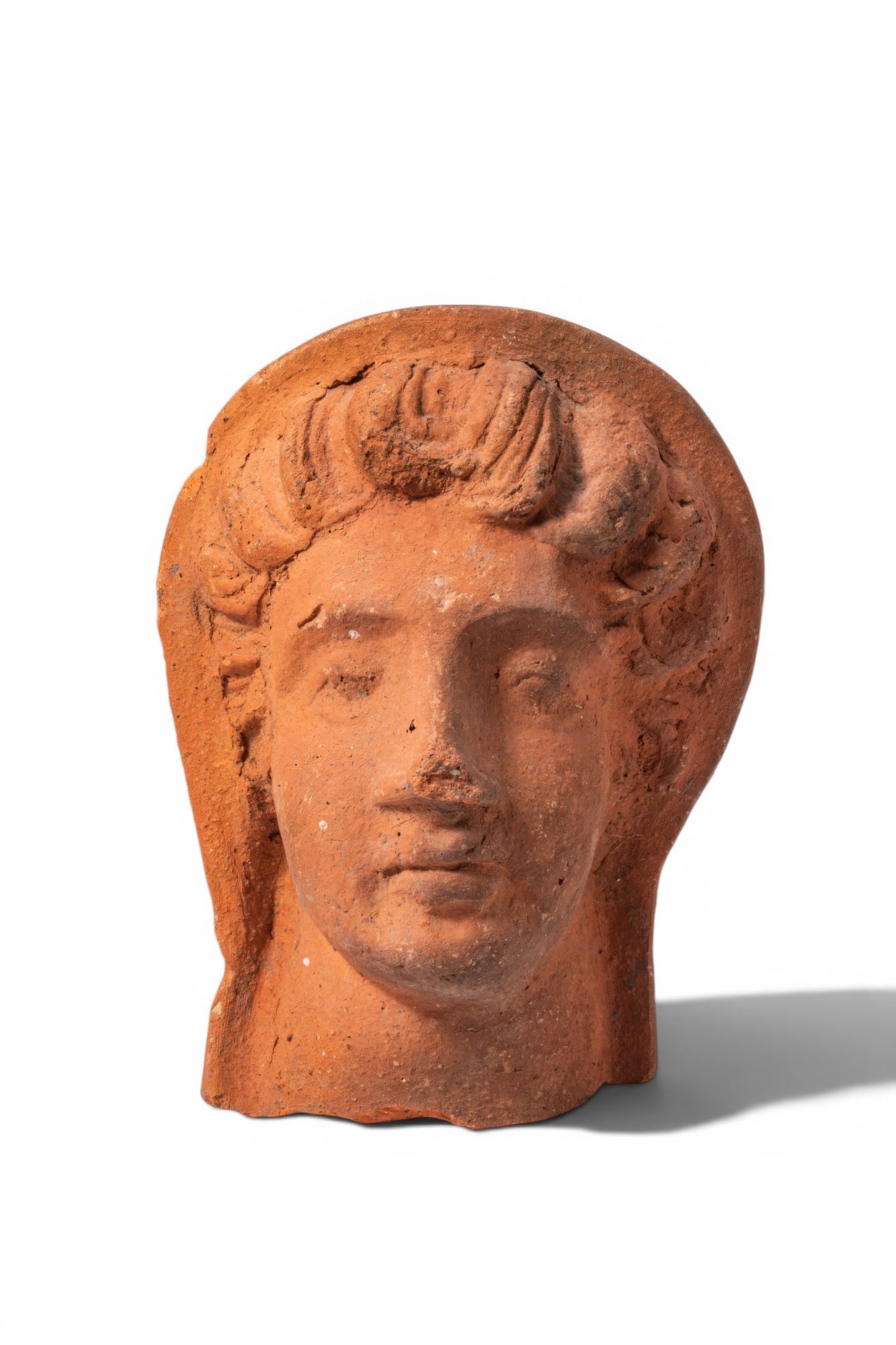 ETRUSCAN TERRACOTTA VOTIVE HEAD OF YOUTH: Ca. 200 - 100 BC. A hollow terracotta votive head of a youth, modelled with an oval face with heavy-lidded eyes, a straight nose and a closed mouth. Short, wavy locks frame the forehead and temples. A