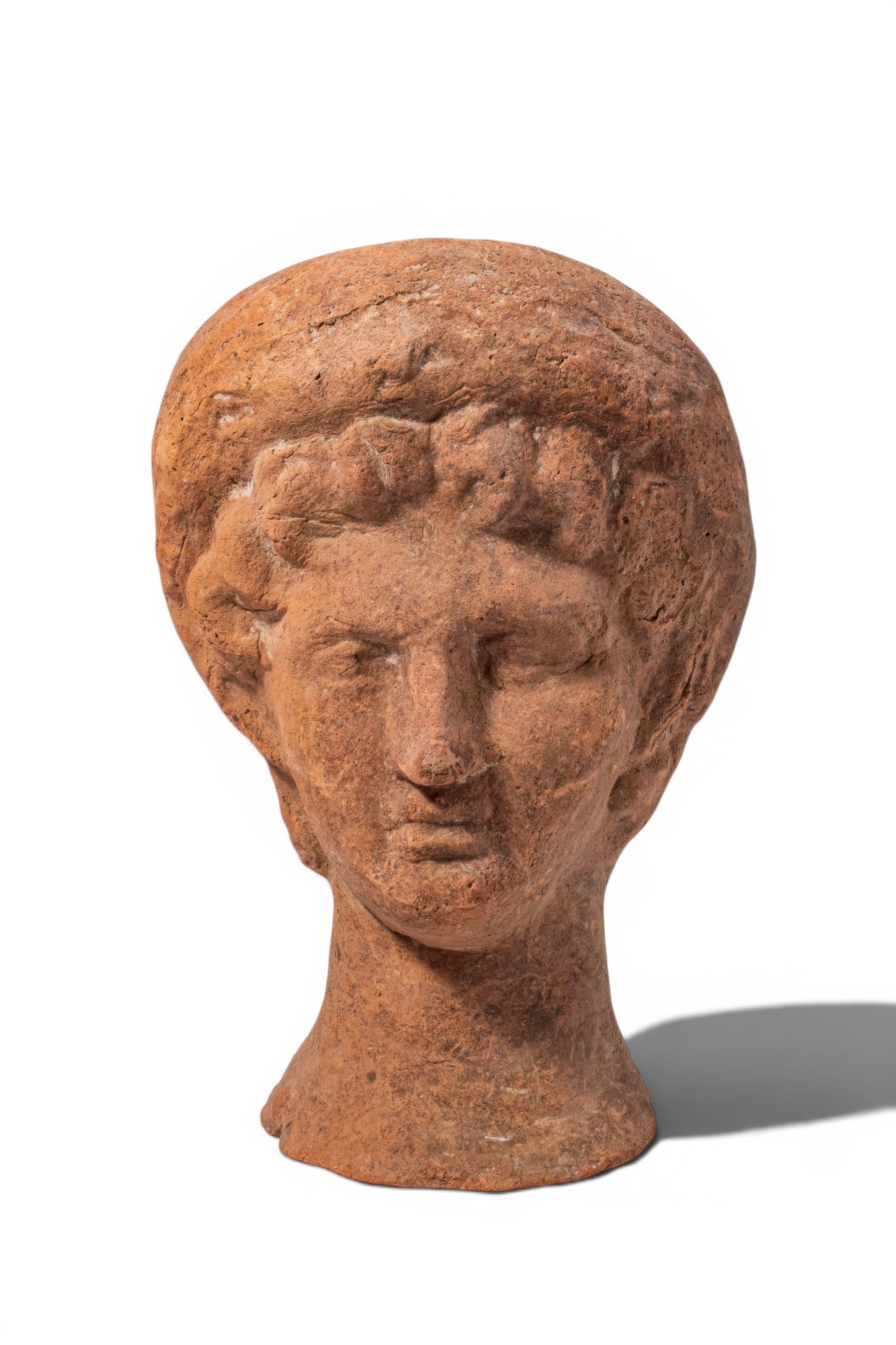 ETRUSCAN TERRACOTTA VOTIVE HEAD OF YOUTH: Ca. 300 - 200 BC. A hollow terracotta votive head of a youth, modelled with an oval face with heavy-lidded eyes, a straight nose and a closed mouth. Short, wavy locks frame the forehead and temples. A