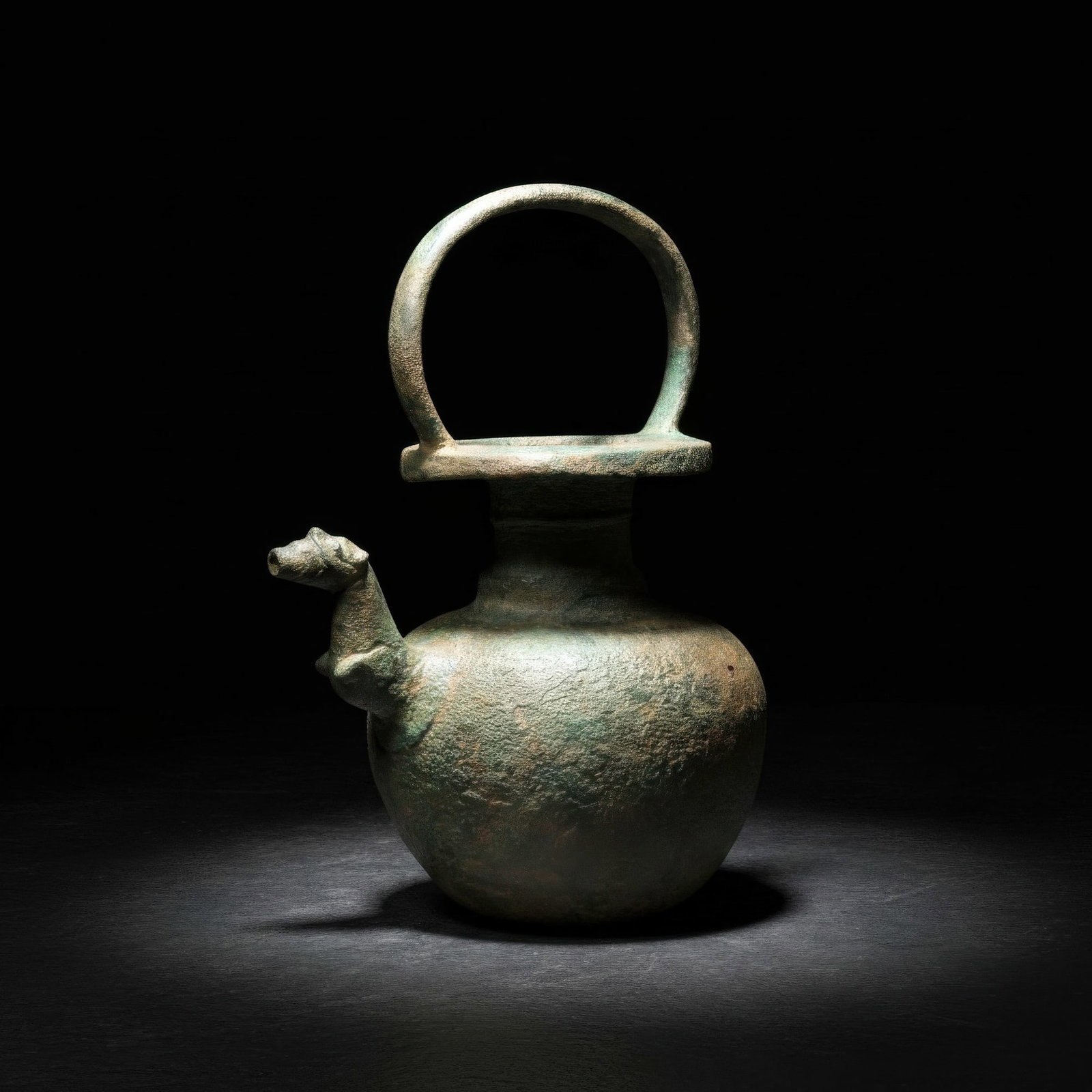 URARTIAN BRONZE EWER WITH DOG PROTOME: Ca. 1000 - 800 BC. A bronze ewer with a globular body and a broad cylindrical neck, with a flat disc mouth surmounted by a large loop handle. A dog-headed spout projects from the shoulder, the narrow 