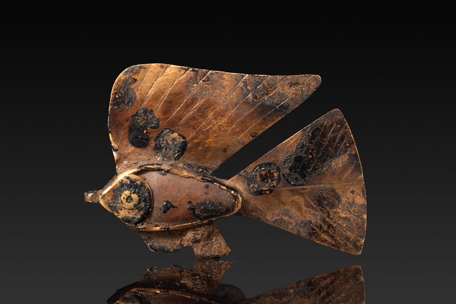 EGYPTIAN GOLD AND STONE FISH AMULET: Middle Kingdom, Ca. 2055 - 1790 BC. A gold amulet in the form of a fish with an oval body inlaid with stone, and broad fin and tail engraved with diagonal hatchings. A small suspension loop projects f