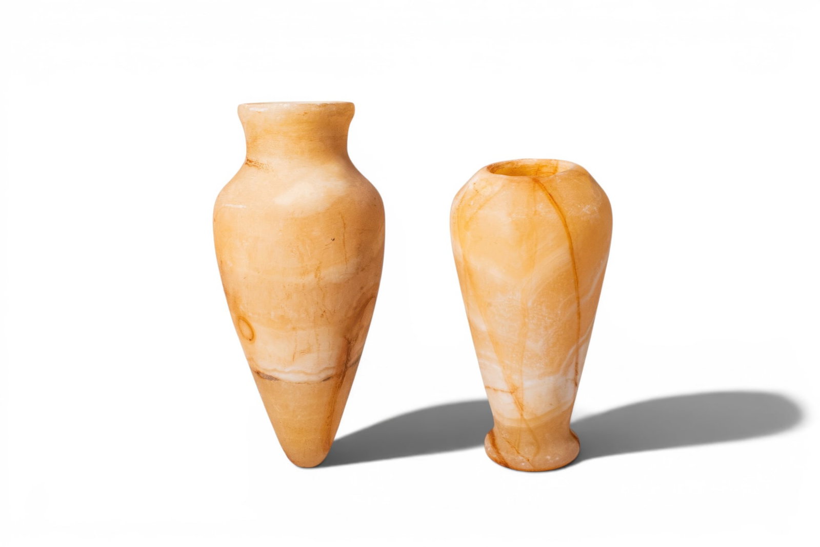 PAIR OF EGYPTIAN ALABASTER JARS: New Kingdom, Ca. 1550 - 1069 BC. A pair of alabaster jars, each with a tall tapering body and a rounded shoulder, the mouth plain; the one on the left with a pointed base, the one on the right on a lo