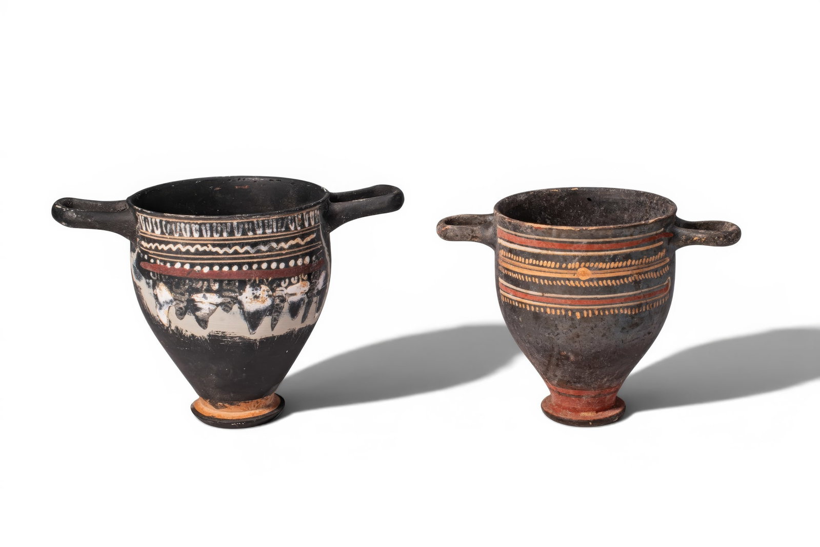 PAIR OF ELEGANT GNATHIAN TERRACOTTA SKYPHOI: Ca. 330 - 270 BC. A pair of terracotta skyphoi, each with a deep tapering cup on a circular foot and a pair of loop handles, decorated in added white, yellow and red; the one on the left painted with 