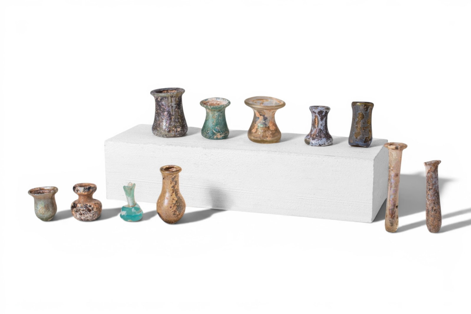 STUNNING COLLECTION OF ROMAN MINIATURE GLASS UNGUENTARIA: Ca. AD 1 - 300. A collection of 11 miniature glass unguentaria, including, from left to right: a tubular bottle with a broad flaring rim; a bottle with a thick rim and globular lower body; a miniature