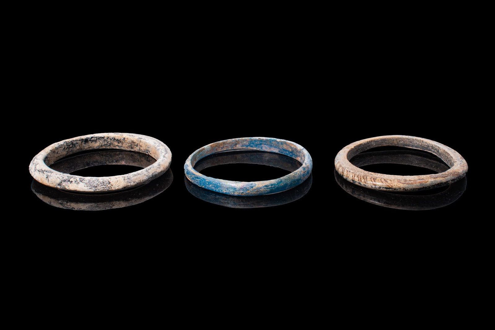 COLLECTION OF ROMAN GLASS BRACELETS: Ca. AD 1 - 300. A collection of three glass bracelets, including, from left to right: black and blue glass bracelets, each with a rounded exterior and a flat interior; and a dark glass bracelet with a