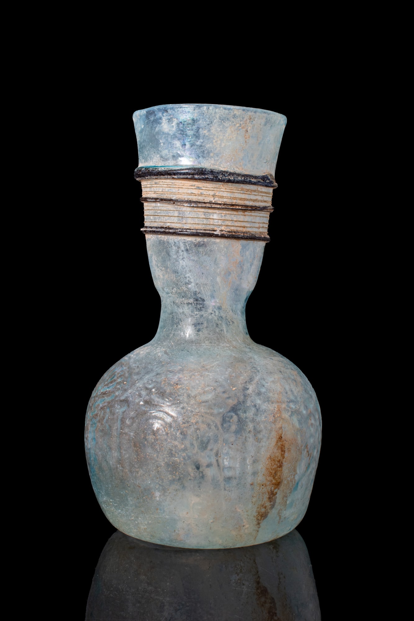 FINE LATE ROMAN GLASS FLASK: Ca. AD 300 - 500. A translucent pale blue glass flask with a rounded mould-blown body and a short, waisted neck below a flaring mouth. The neck is encircled by applied dark blue rigaree bands framing 