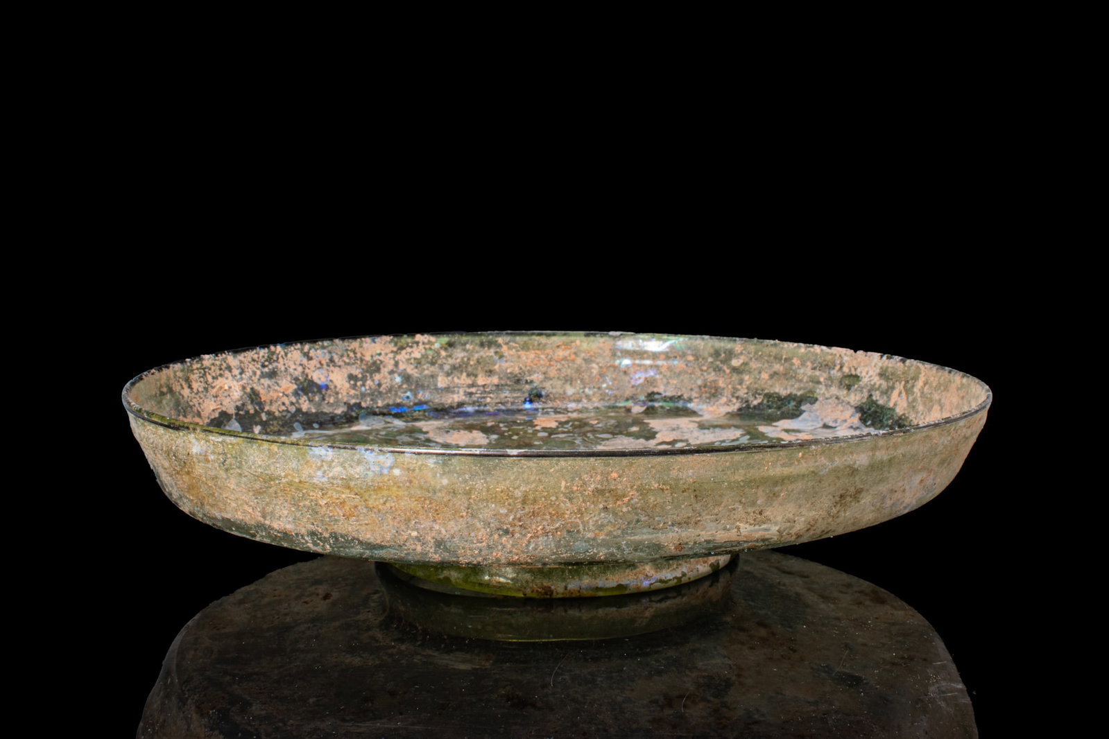 ROMAN GLASS FOOTED DISH: Ca. AD 300 - 400. A free-blown translucent olive green glass dish of a broad shallow form with a curving wall and a plain rounded rim, set above a low ring foot. For similar see: The Metropolitan Muse
