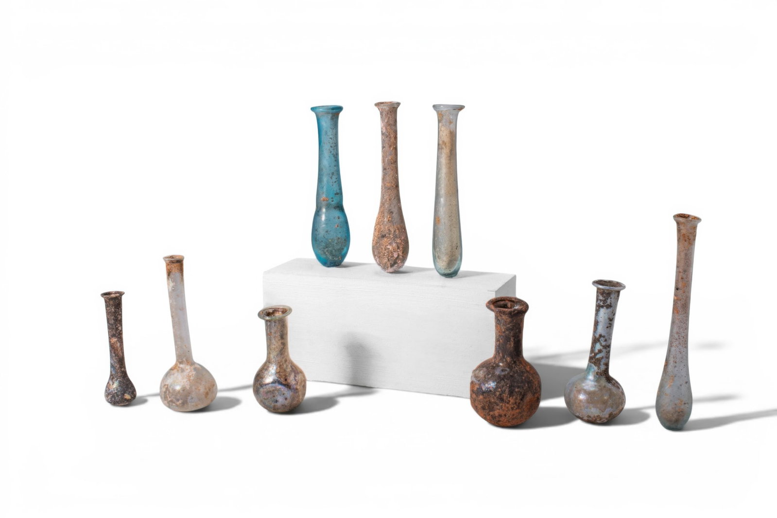 LARGE COLLECTION OF ROMAN GLASS UNGUENTARIA: Ca. AD 1 - 400. A collection of nine glass unguentaria, including, from left to right: a bottle with a small piriform body and a tall cylindrical neck; a bottle with a broad globular body and a long n