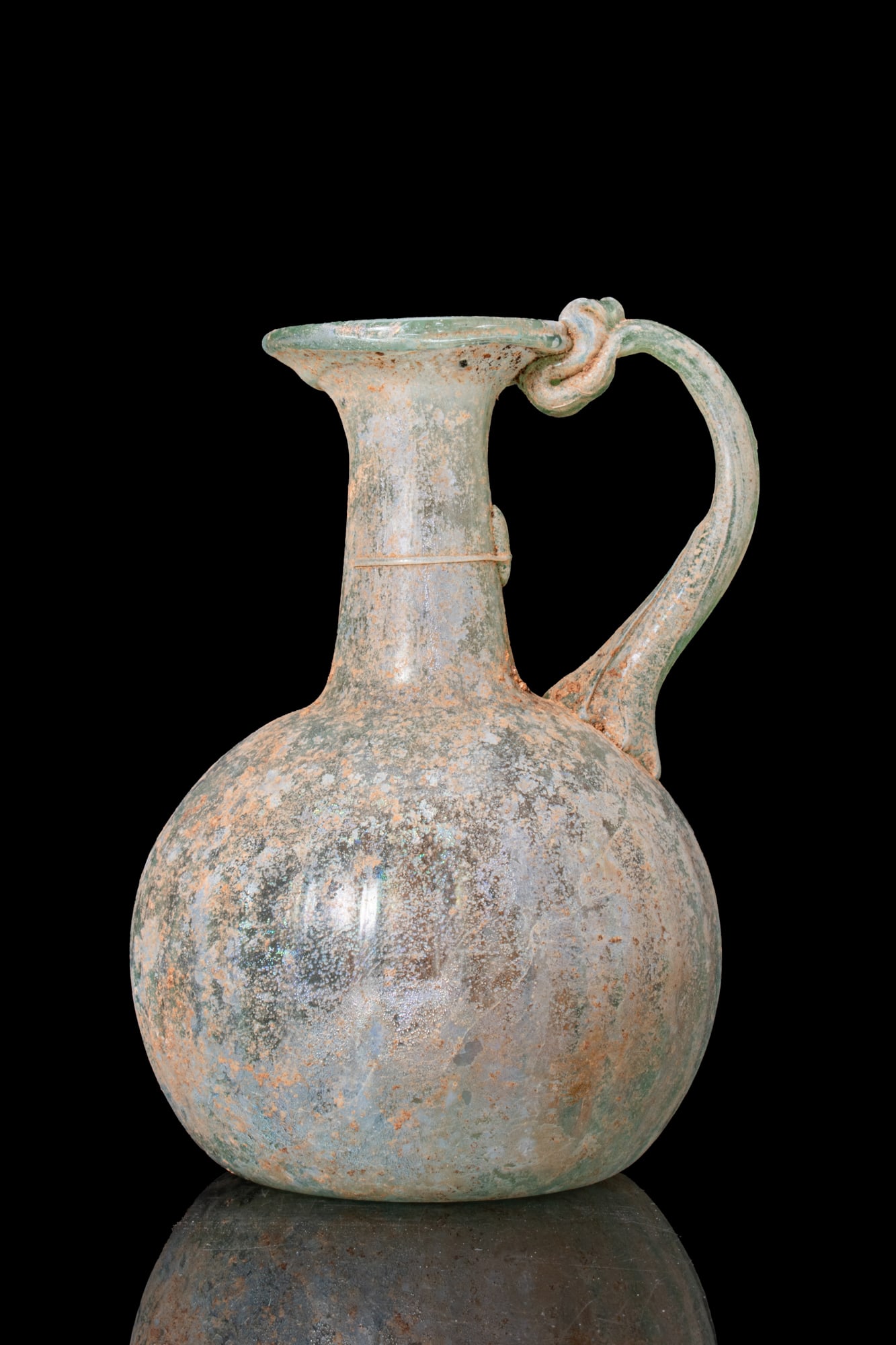 LATE ROMAN GLASS JUG: Ca. AD 300 - 400. A translucent pale green glass jug with an onion-shaped body decorated with vertical fluting, a tall cylindrical neck widening to a funnel mouth, and a strap handle attached between 