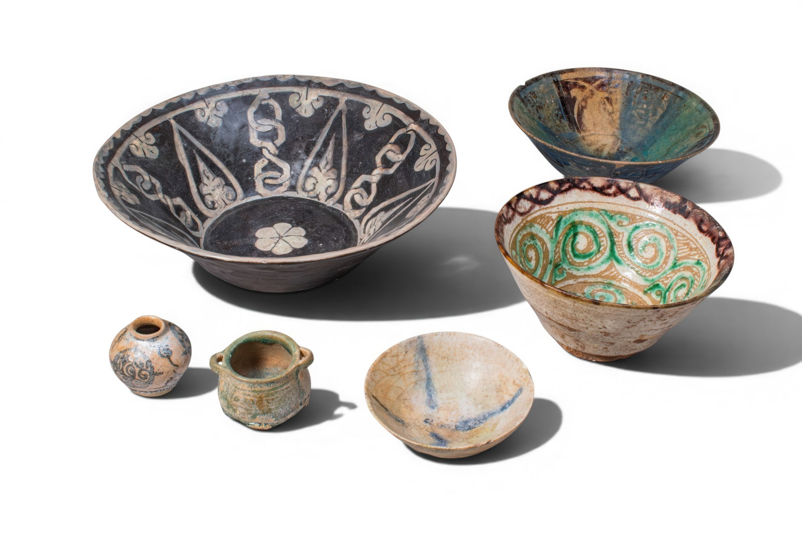 COLLECTION OF KASHAN GLAZED VESSELS WITH VEGETAL AND FLORAL DECORATION: Ca. AD 1200 - 1500. A collection of six vessels, including, from left to right: a globular jar with a short neck and narrow mouth, painted with scrolling motifs in dark blue on a pale ground; a miniat