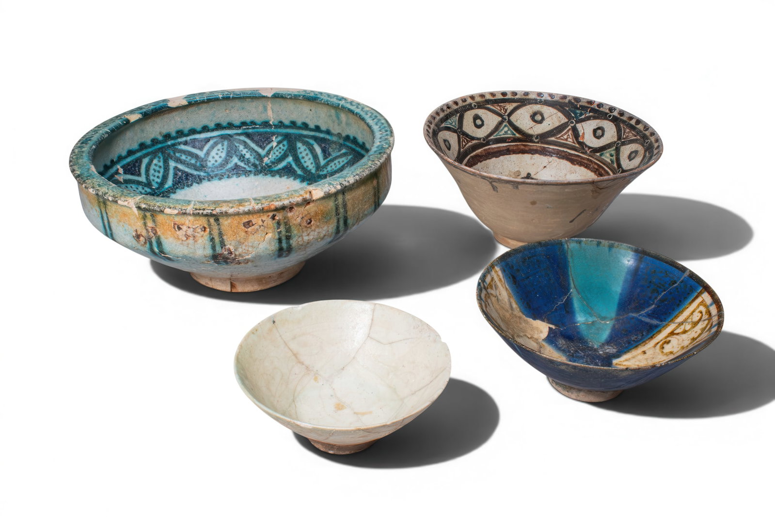 COLLECTION OF KASHAN GLAZED BOWLS WITH VEGETAL AND FLORAL DECORATION: Ca. AD 1000 - 1400. A collection of four pottery bowls, including, from left to right: a large deep bowl on a short foot, painted in turquoise with concentric bands of stylised leaf ornament and a cen
