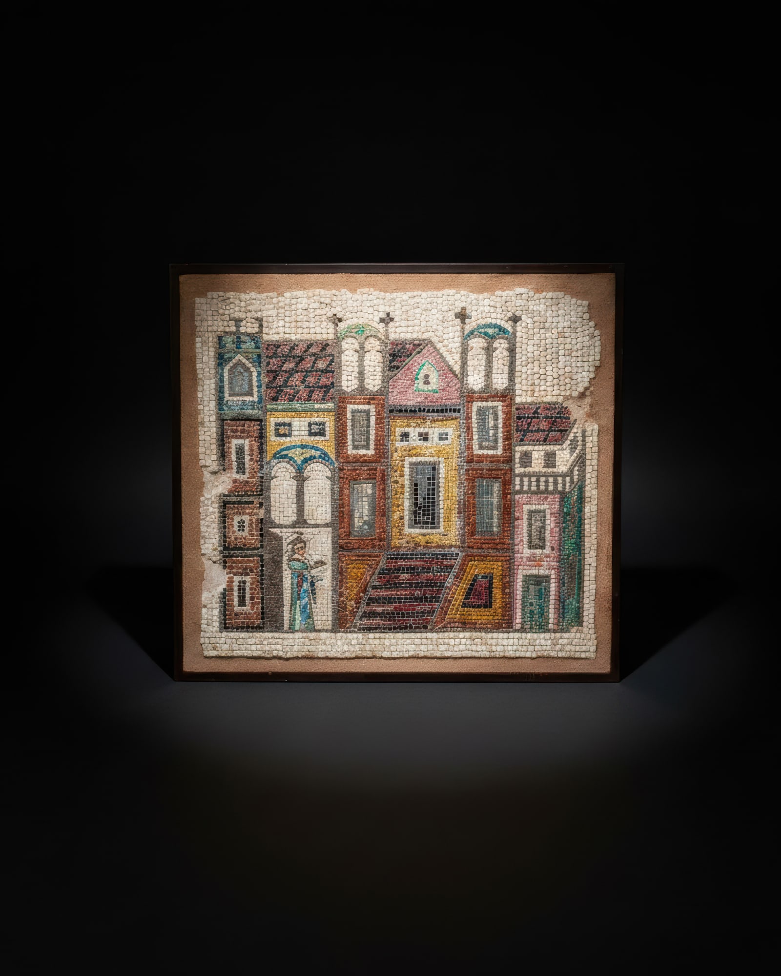 MOSAIC PANEL WITH ARCHITECTURAL SCENE: . A framed mosaic panel composed of ancient tesserae, depicting a dense architectural facade with towers, arched windows, gabled roofs, stairways, and doorways. The buildings are arranged in tiers acr