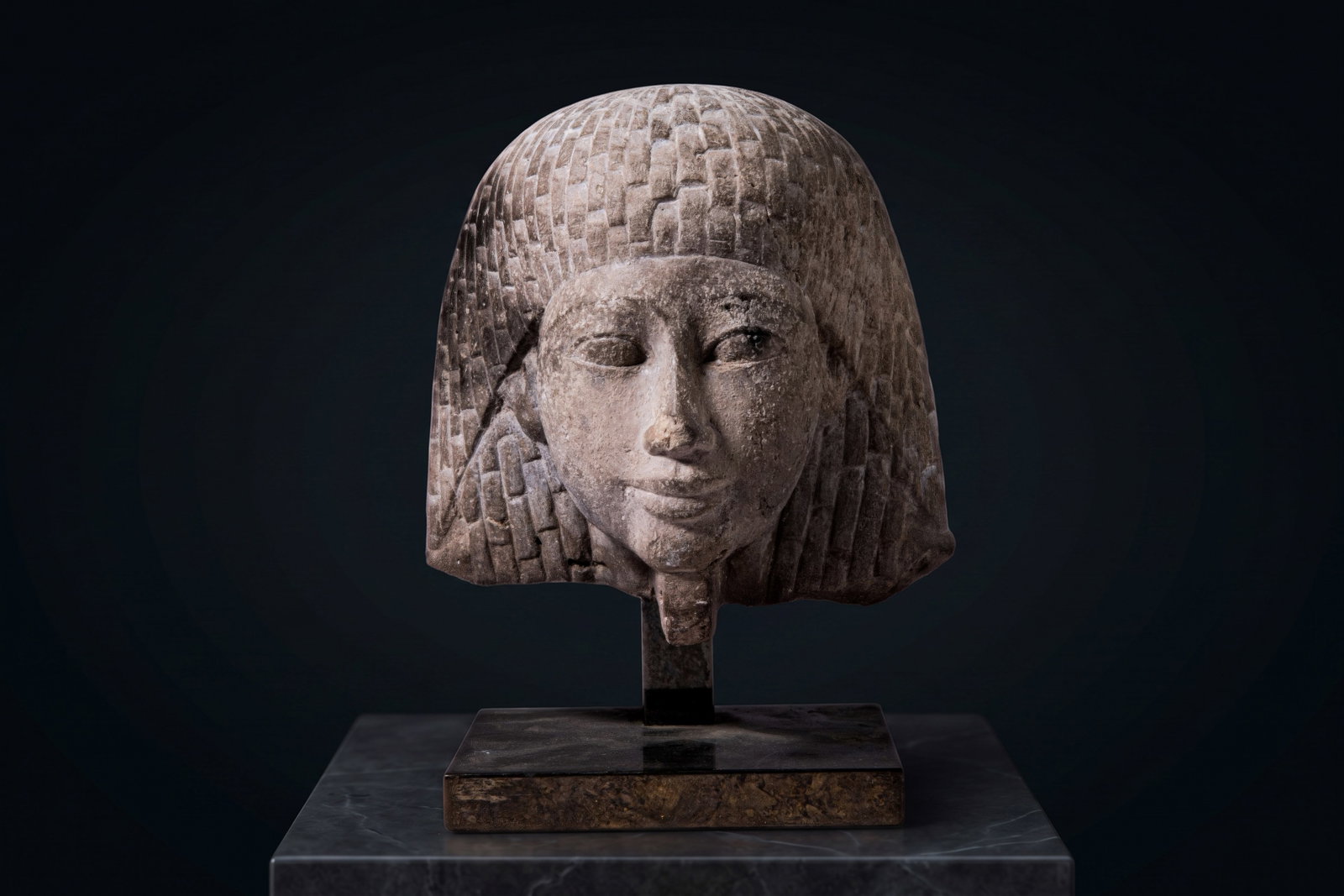 EGYPTIAN OLD KINGDOM STONE HEAD OF PRINCE: New Kingdom, Ca. 1550 - 1069 BC. A stone head of a prince with a broad face, almond-shaped eyes, a straight nose and softly modelled lips. The head is framed by a voluminous curled wig extending to th