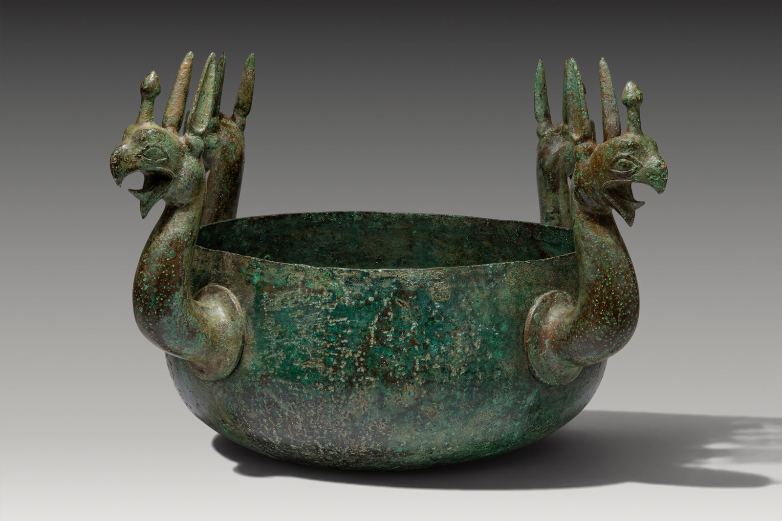 GREEK BRONZE CAULDRON WITH GRIFFIN PROTOMES: Ca. 700 - 600 BC. A bronze cauldron with a deep rounded bowl and a plain upright rim. Four griffin protomes project at regular intervals around the exterior, each with a long curved neck, an open hook