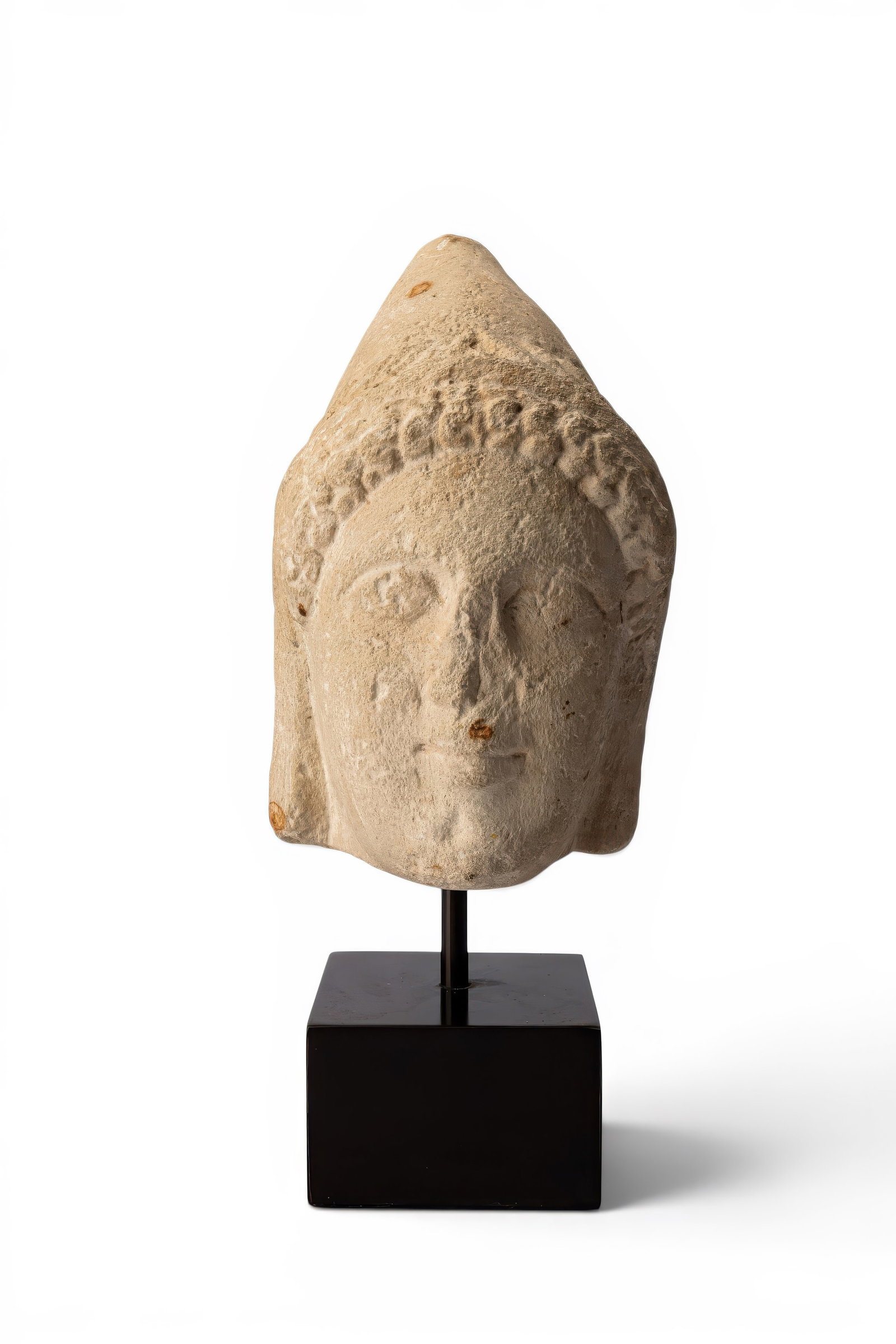 CYPRIOT MARBLE HEAD OF A MAN WEARING A CONICAL CAP: Ca. 550 - 500 BC. A marble head of a man wearing a tall conical cap, its lower edge bordered by a row of tongues. The full face is carved with heavy-lidded almond-shaped eyes, a strong straight nose a