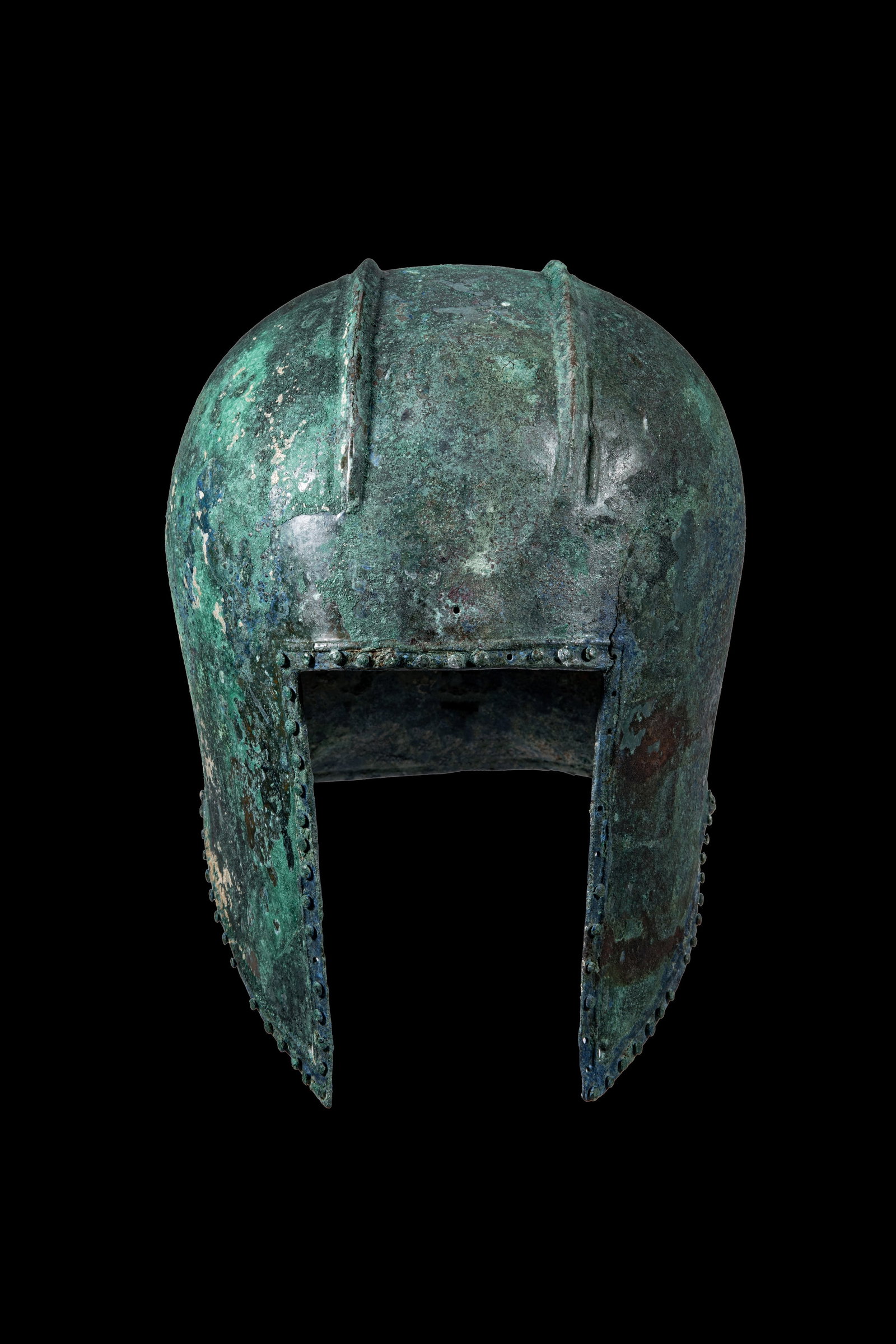 ILLYRIAN BRONZE HELMET: Ca. 500 BC. A bronze helmet with a rounded skull and a rectangular face opening framed by cheekpieces tapering downwards. Two pronounced longitudinal ribs extend over the crown. Rows of rivets border 