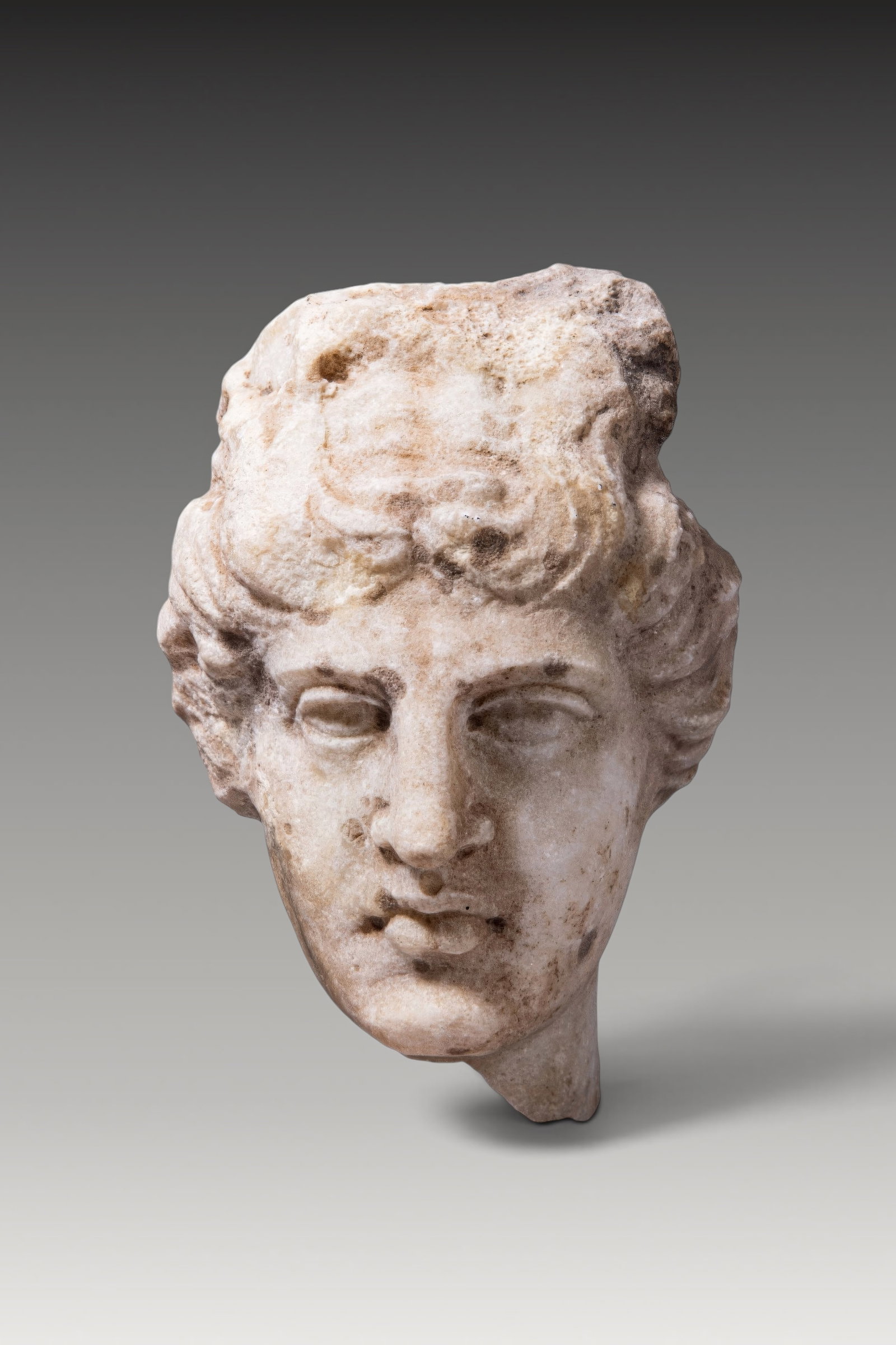 EASTERN ROMAN MARBLE HEAD OF APOLLO BELVEDERE TYPE: Ca. AD 200. A marble head of Apollo of Belvedere type, with the hair arranged in thick, deeply carved locks. The curls are swept back from the forehead and gathered into a central knot, with further l