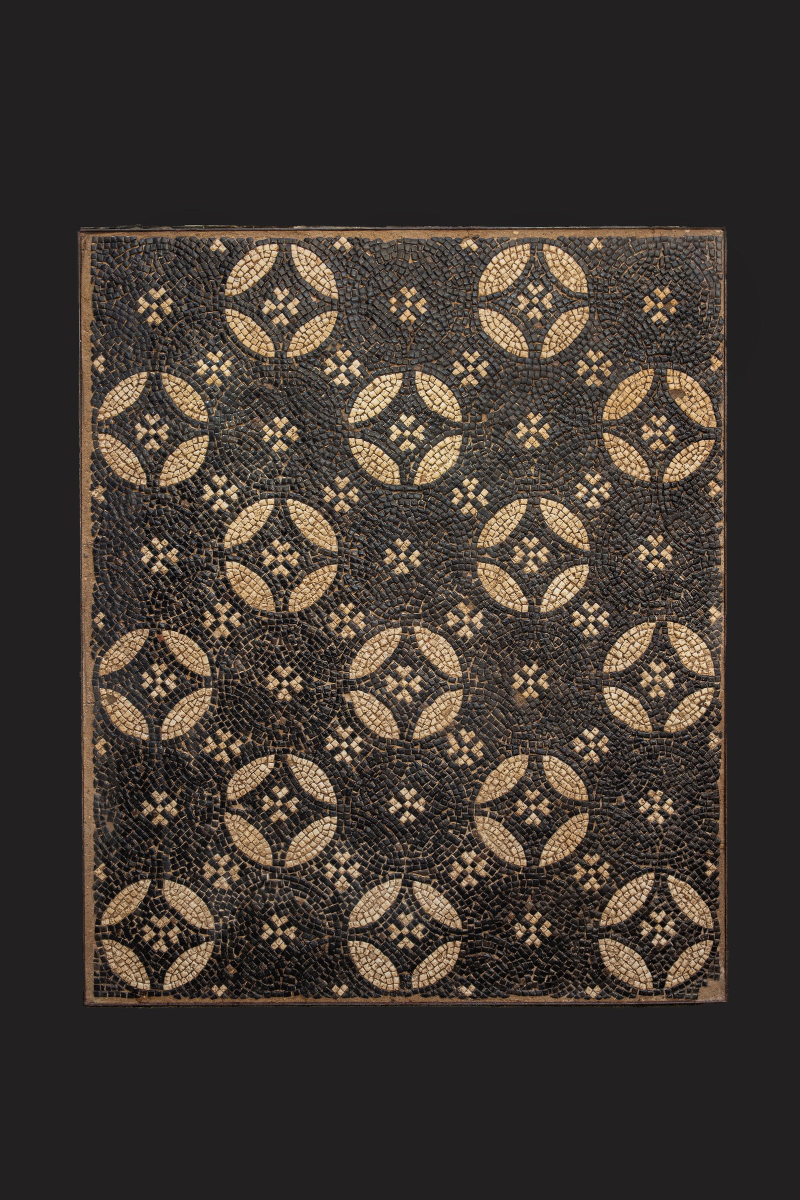 ROMAN MOSAIC PANEL WITH GEOMETRIC MOTIFS, OSTIA STYLE: Ca. AD 175 - 200. A rectangular mosaic panel composed of black, cream, and ochre tesserae. The field is arranged with regularly spaced circular geometric medallions. Each medallion contains four inwar