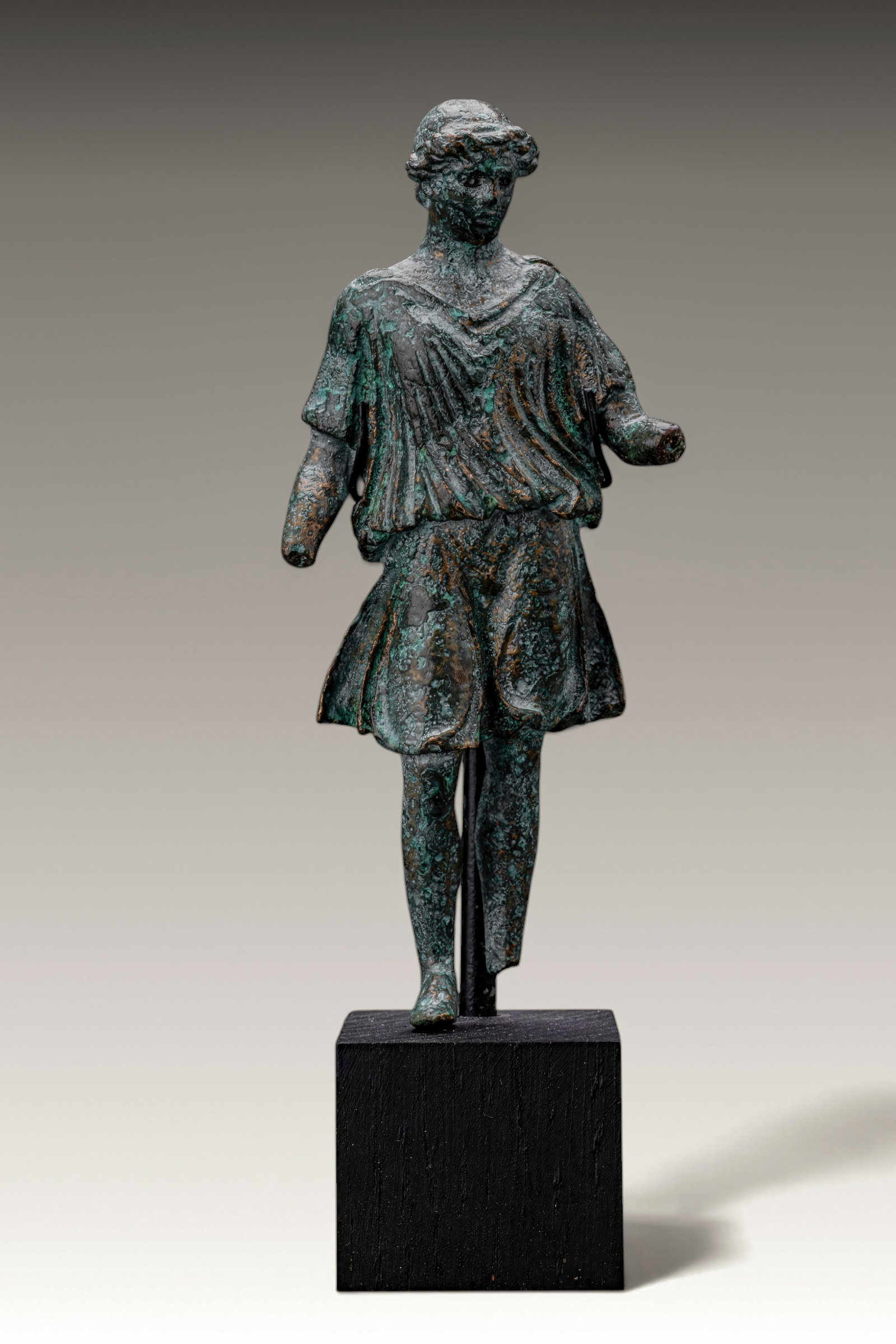 ROMAN BRONZE LAR FIGURINE: Ca. AD 1 - 300 . A bronze figurine of a youthful Lar standing with the right leg advanced, dressed in a short belted tunic falling in deep folds. Thick curling hair frames the face. The left arm is ex