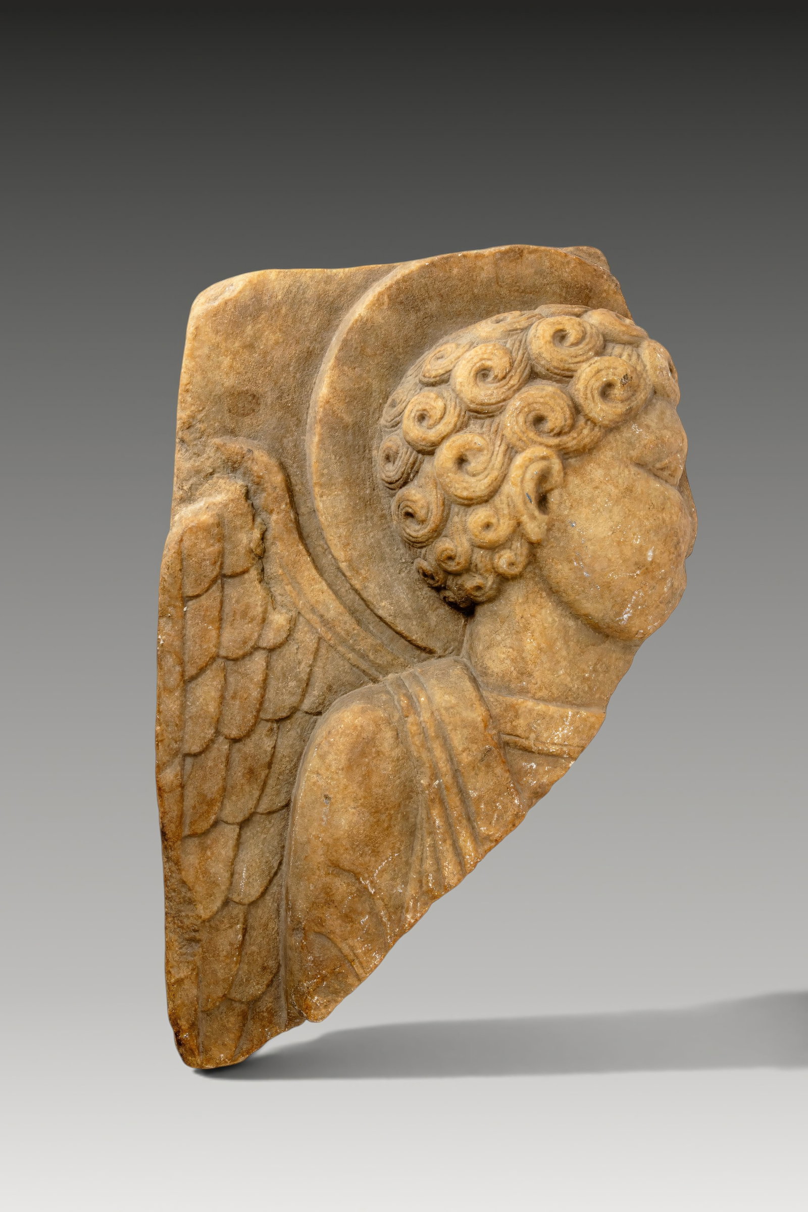 BYZANTINE MARBLE RELIEF OF AN ANGEL: Ca. AD 1200. A marble relief depicting an angel in profile to the right, with a circular halo behind the head. Rows of tightly curled hair frame the face and descend to the nape. A feathered wing exte