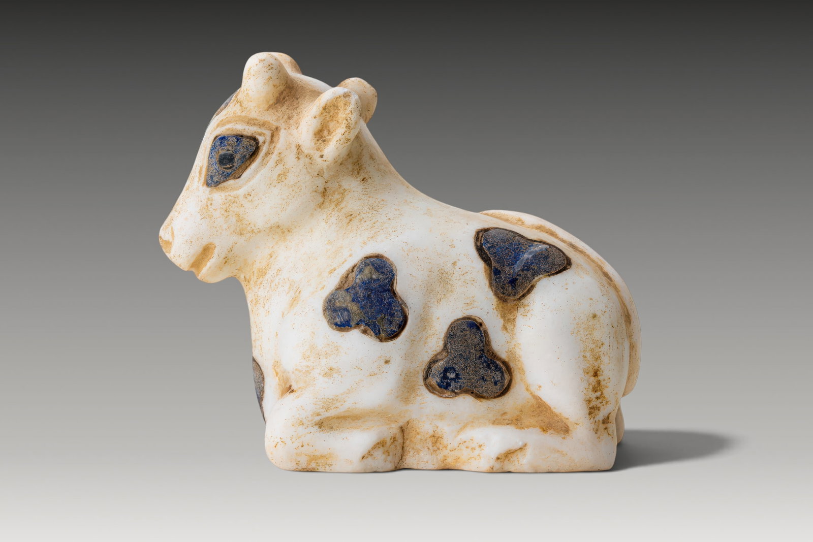 MESOPOTAMIAN MARBLE AND LAPIS LAZULI INLAID BULL OF HEAVEN IDOL: Ca. 3500 - 3000 BC. A recumbent marble bull, carved with the legs folded beneath the body, the head raised and turned slightly to one side. The eyes, ears, and a series of trefoil-shaped patches acros